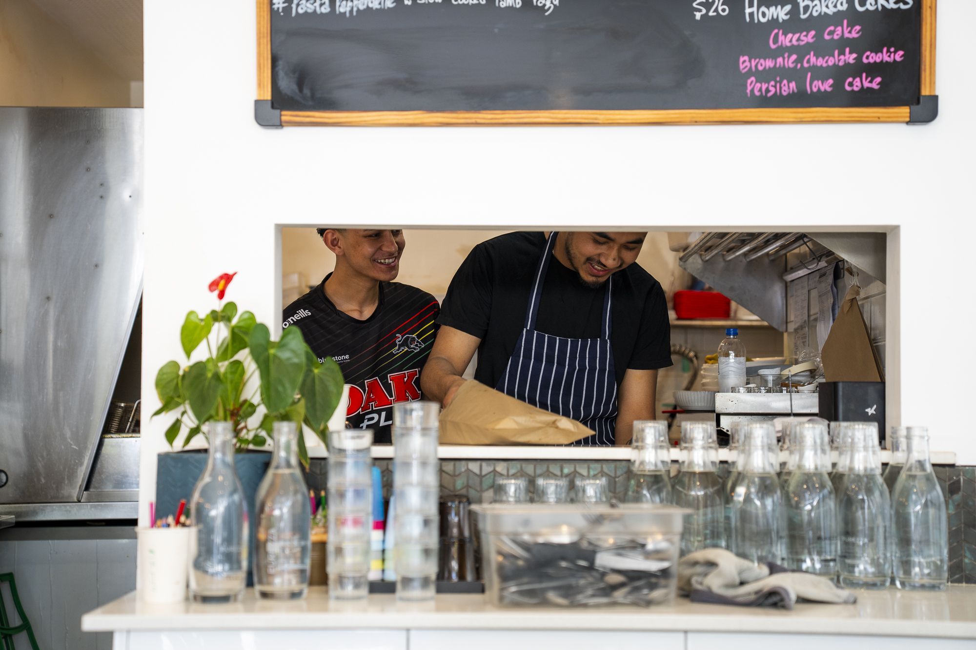 Two people smiling and preparing food in a kitchen, viewed through a serving window. There are bottles and potted plants on the counter.