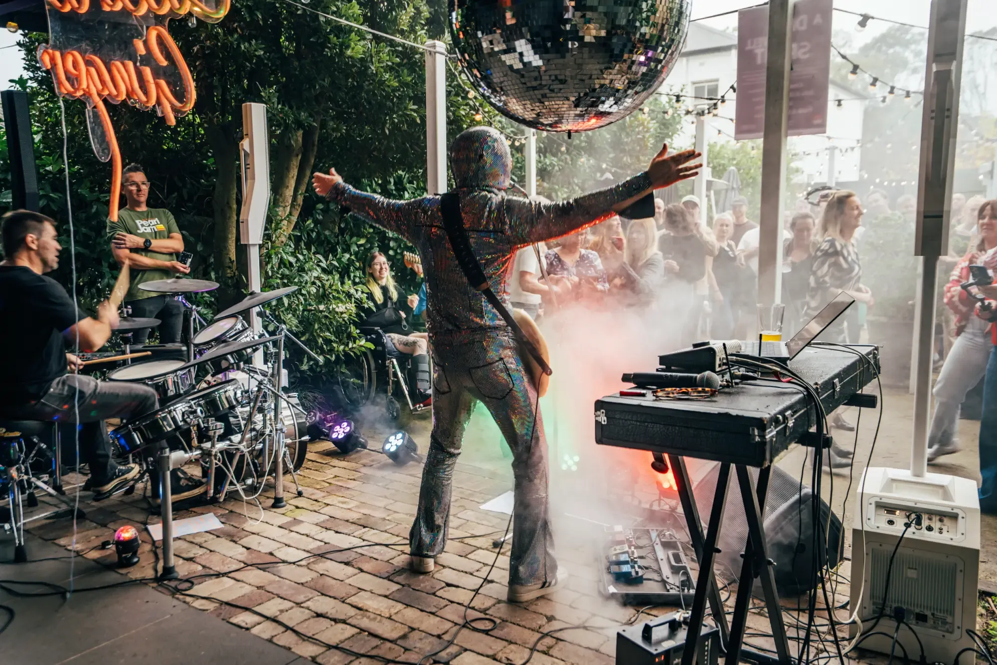 Musician on stage with arms raised, a disco ball overhead, surrounded by instruments and audience under an outdoor covered area.