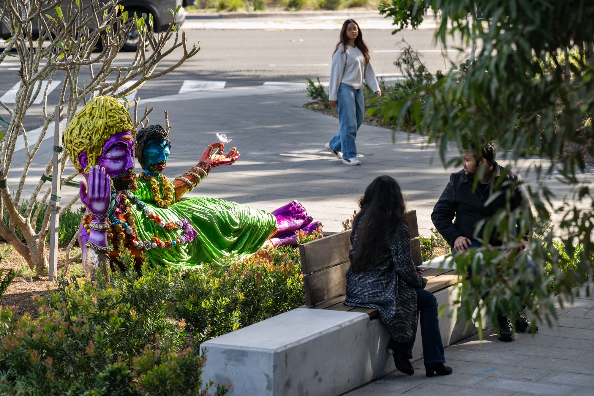 Vibrant multi-headed reclining sculpture in purple, green, and gold with beaded adornments, installed in a landscaped public space where two people sit on a bench and another person walks past.