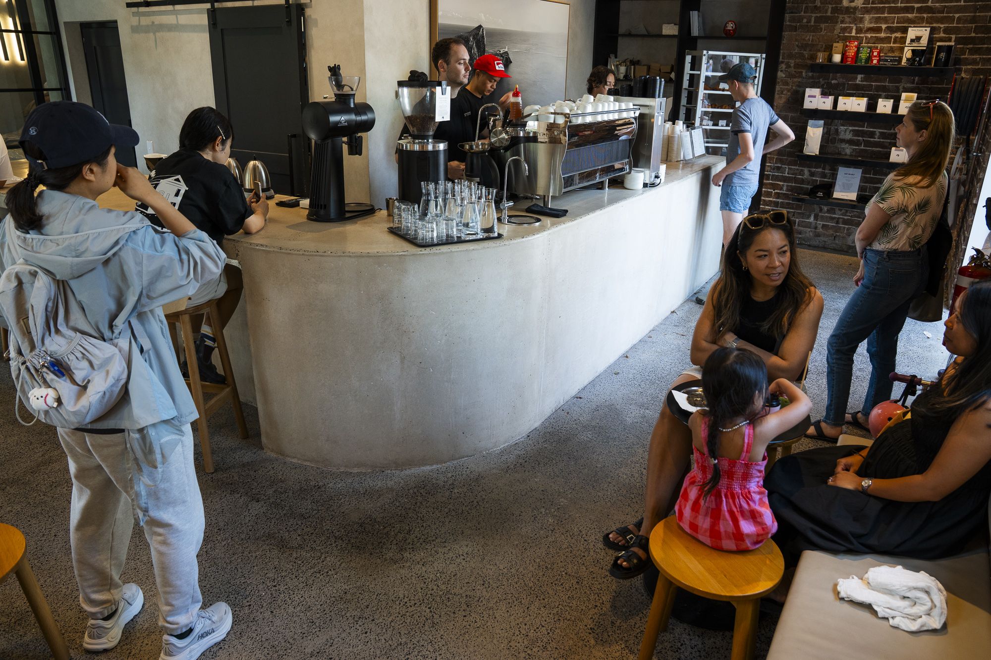Customers in a modern café, some ordering at the counter, while others sit and converse at a small table. Baristas are preparing drinks.
