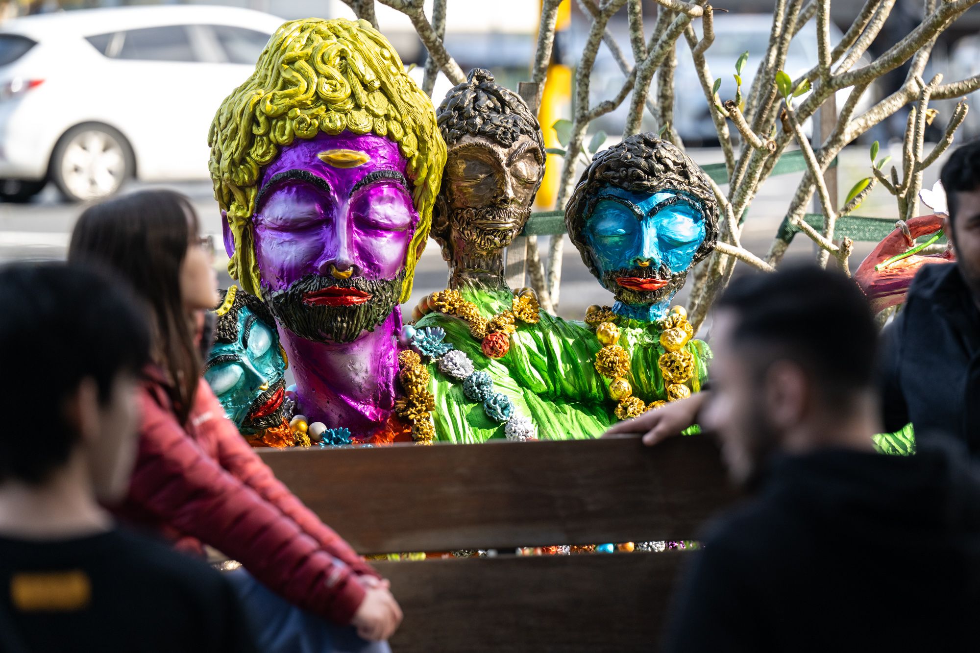 Close-up of a vibrant multi-headed reclining sculpture in purple, blue, green, and gold, adorned with beaded necklaces, with people sitting on a bench in the foreground.