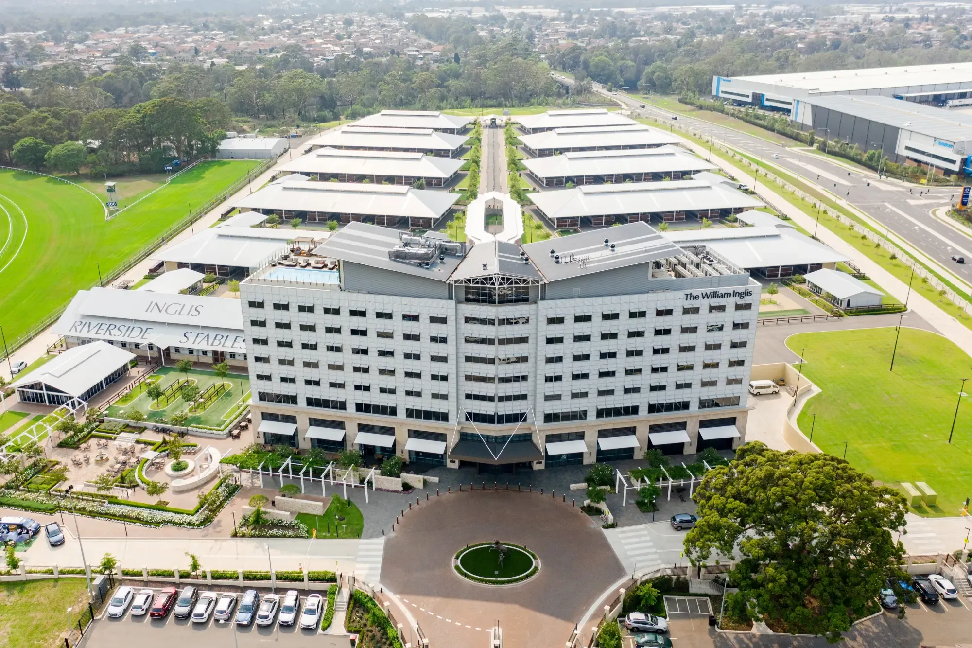 Aerial view of a large, multi-story building surrounded by green fields, parking areas, and adjacent structures, labeled "The William Inglis."