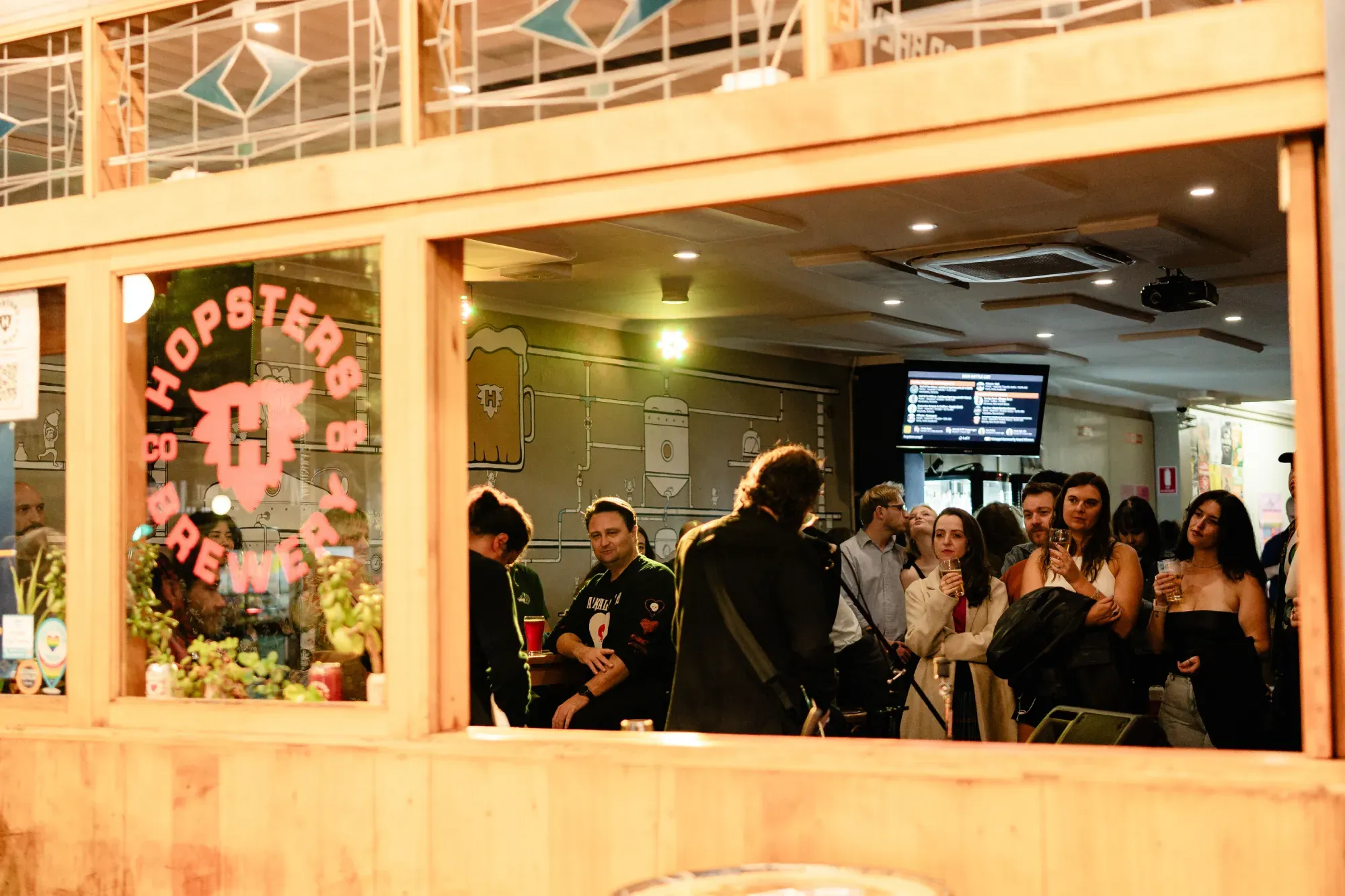 People gathered inside a brewery, with a wooden window frame reflecting "Hopsters Co-Op Brewery." Indoor lighting and a menu screen visible.