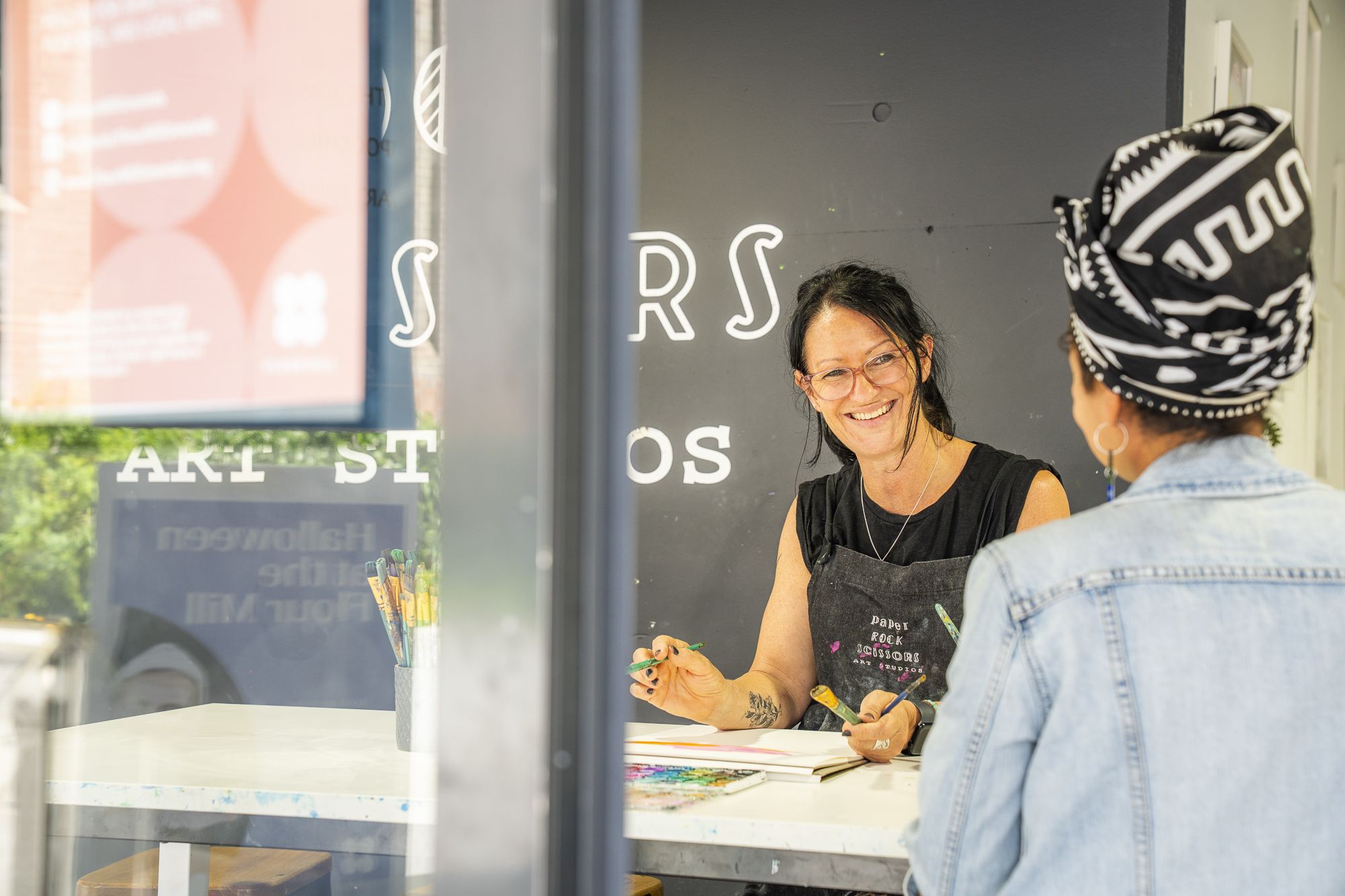 A smiling woman in glasses, wearing an apron, sits at a table with art supplies, interacting with another person in a headwrap and denim jacket.