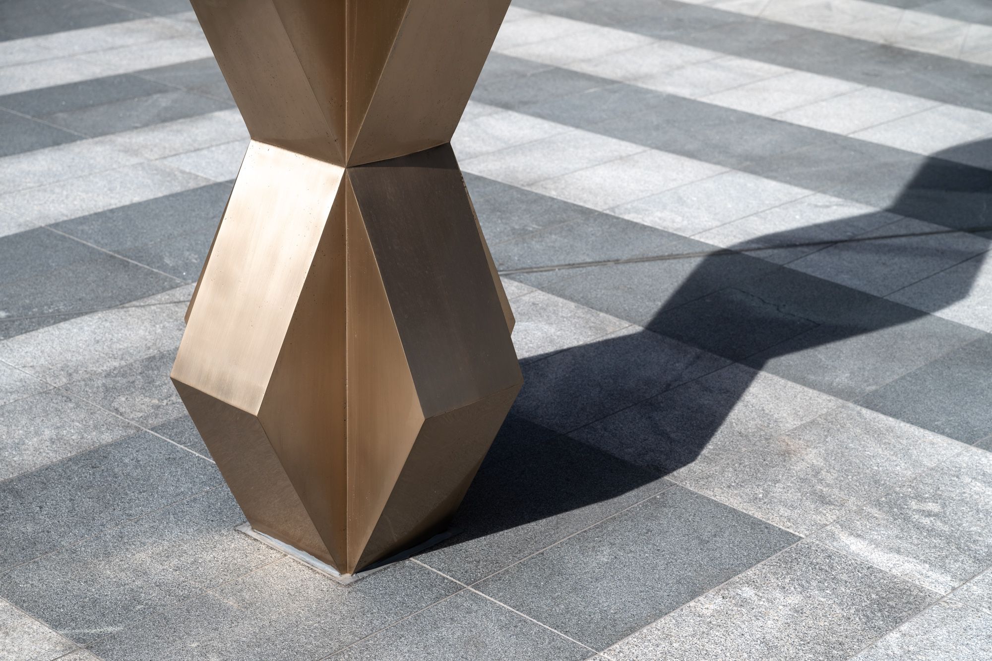 Close-up of the bronze base of a faceted geometric sculpture casting a sharp shadow across a grey stone tiled pavement.