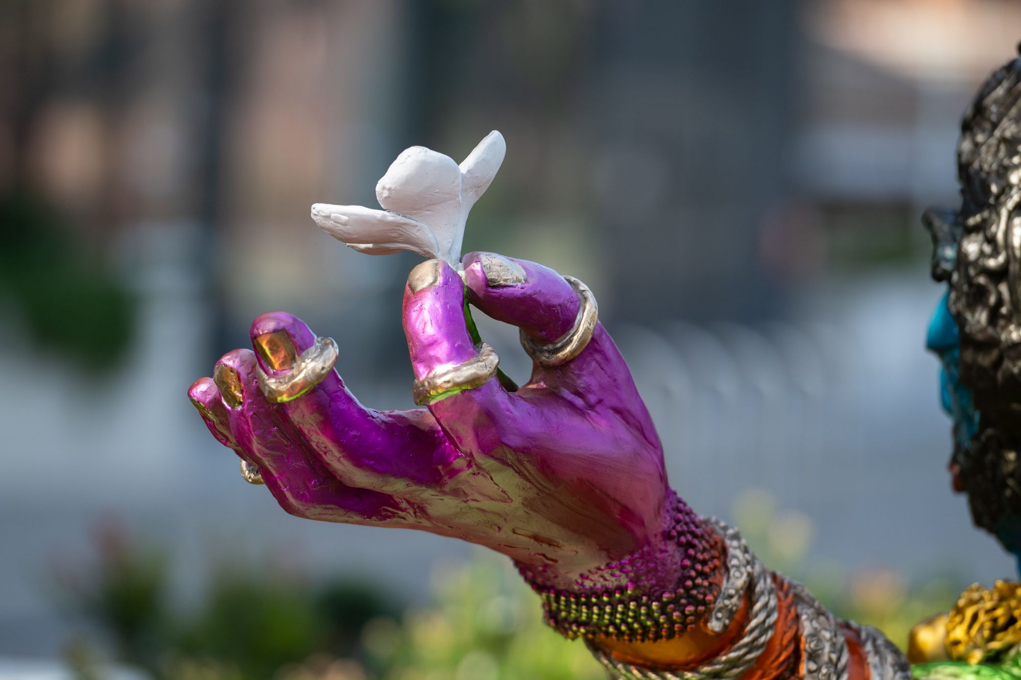 Close-up of a vibrant purple sculpted hand adorned with gold rings and bracelets, delicately holding a white flower.