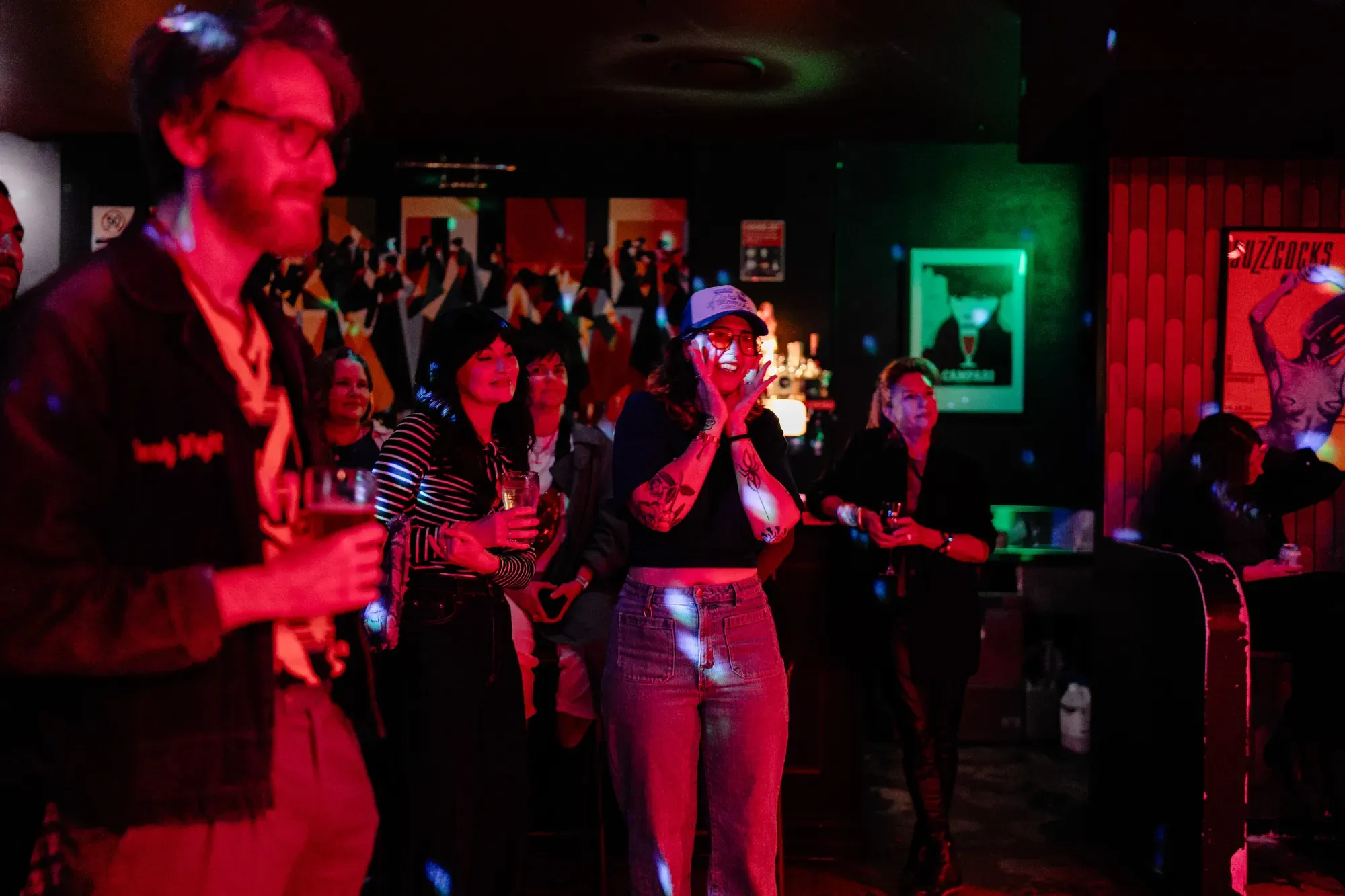 A group of people in a dimly lit bar, some holding drinks, with colorful lights and posters on the walls. One person looks surprised.