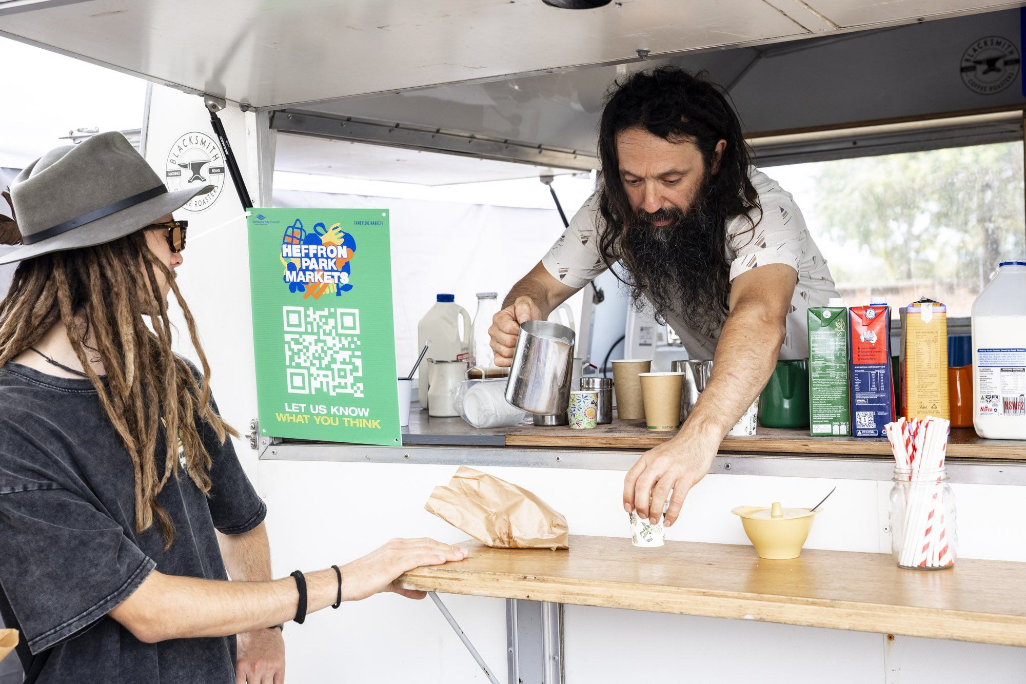 Barista with long hair and beard serves coffee to a customer in a hat at an outdoor market stall. Various milk cartons are visible.