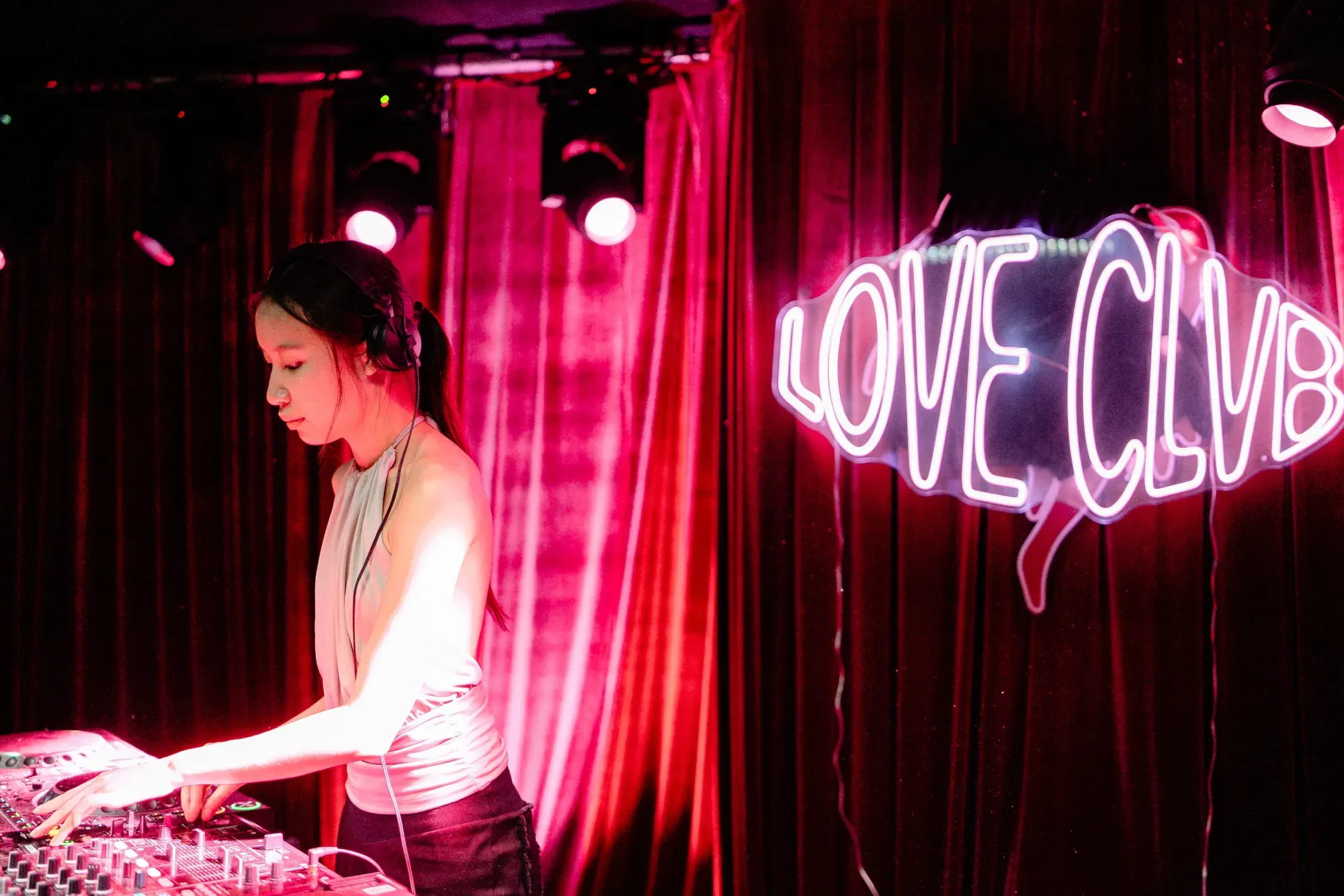 DJ in a club with headphones, mixing music on a DJ console, under pink lights. Neon sign reads "LOVE CLUB" in the background.