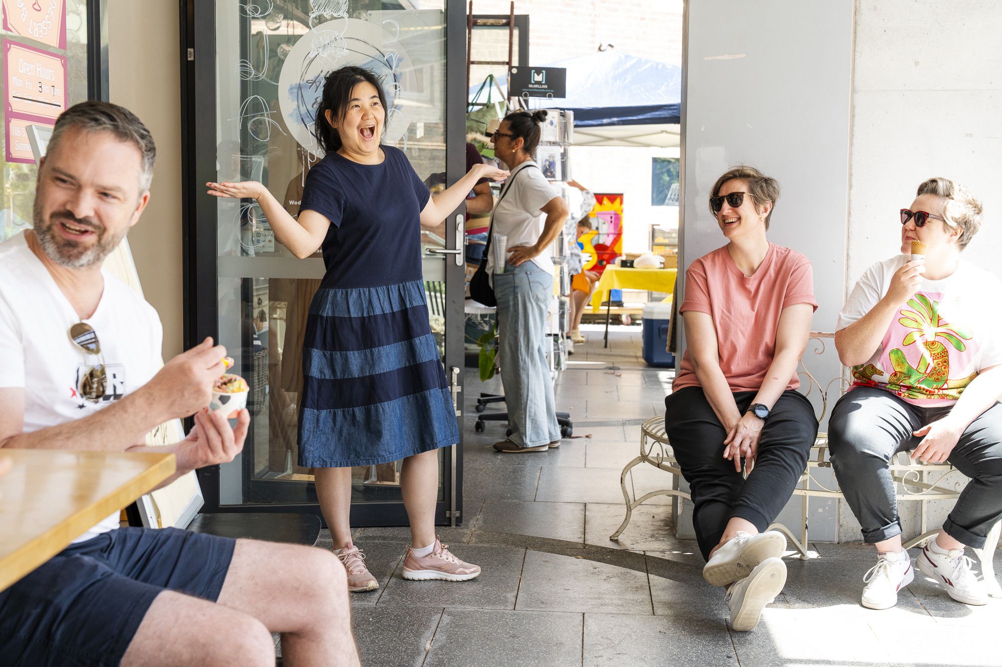 A woman joyfully gestures while three people seated nearby smile and observe. One person is eating ice cream. Outdoor setting, sunny day.