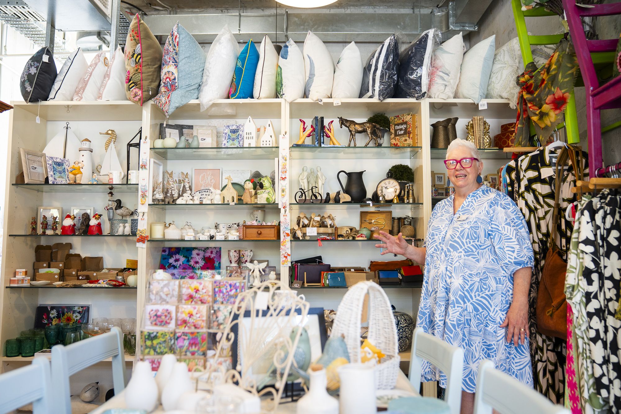 A woman in a blue patterned dress stands in a shop filled with decorative items and colorful pillows on shelves.