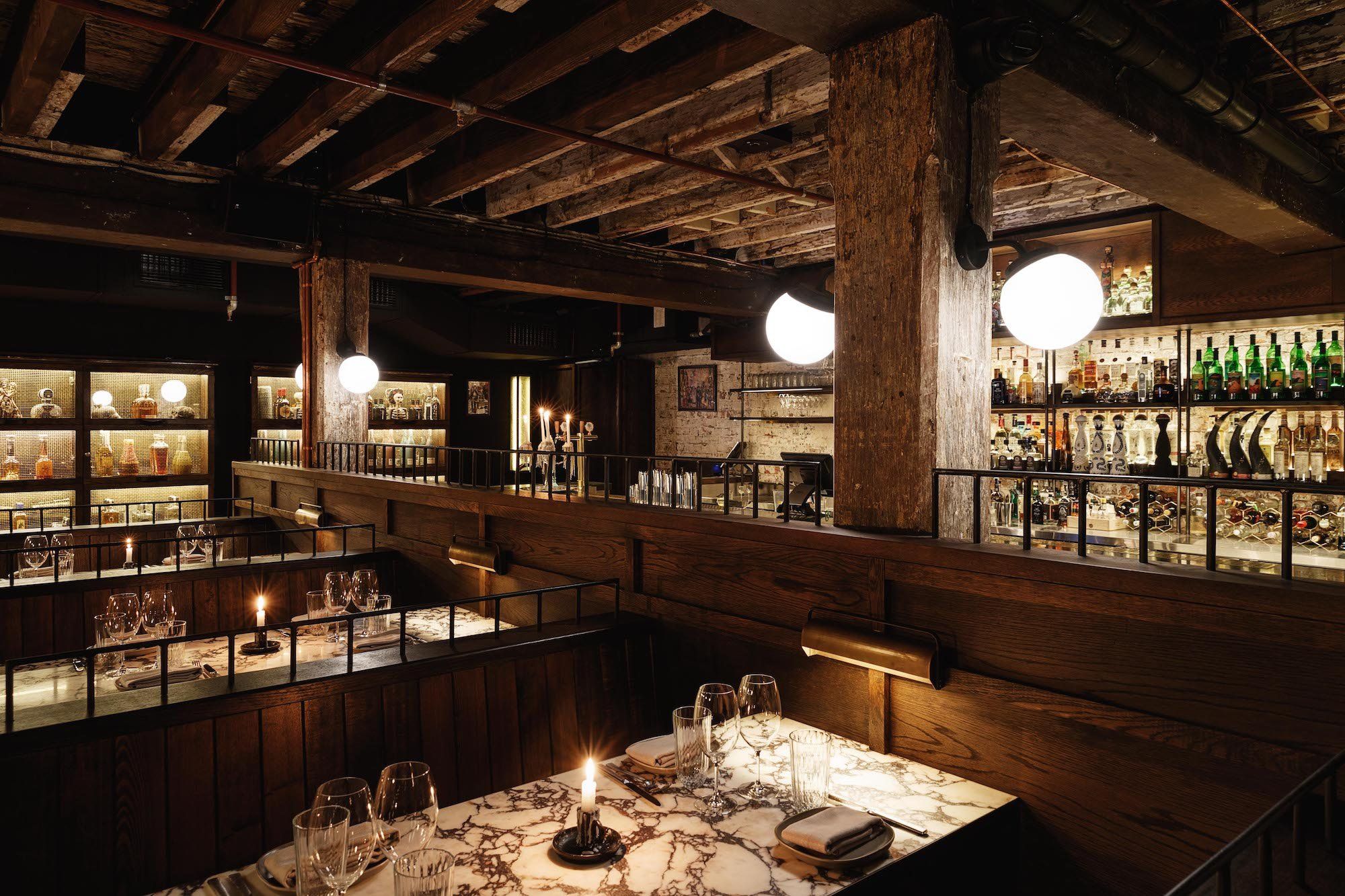 Cozy restaurant interior with wooden beams, dim lighting, and candlelit tables. Shelves stocked with bottles, creating a warm, inviting ambiance.
