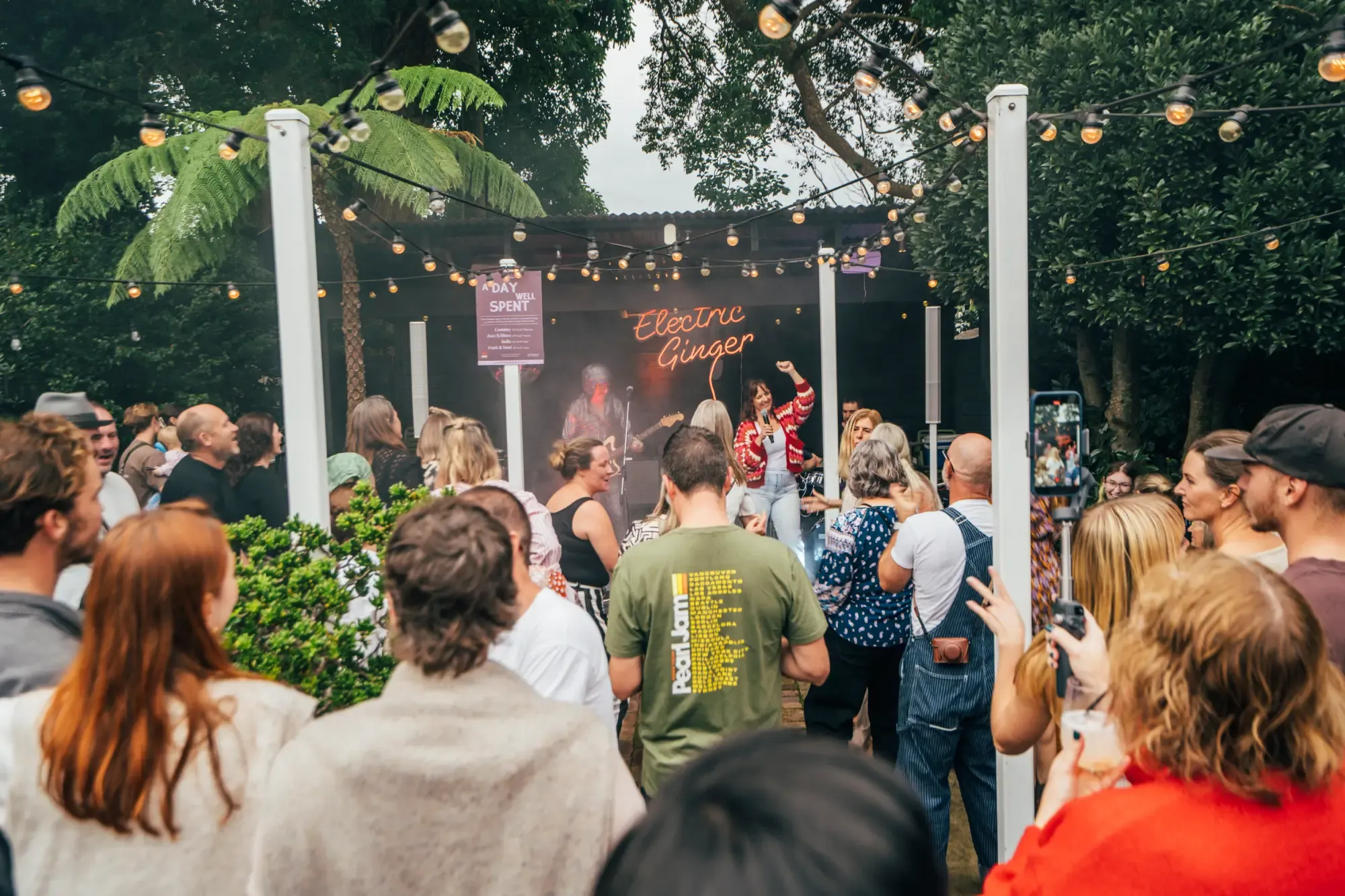 Outdoor concert scene with a crowd watching a band perform on a small stage surrounded by string lights and trees.