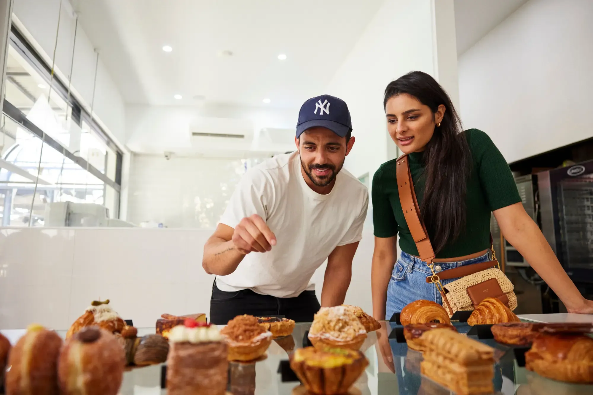 A man and woman eagerly look at a display of various pastries on a glass counter in a bright bakery.