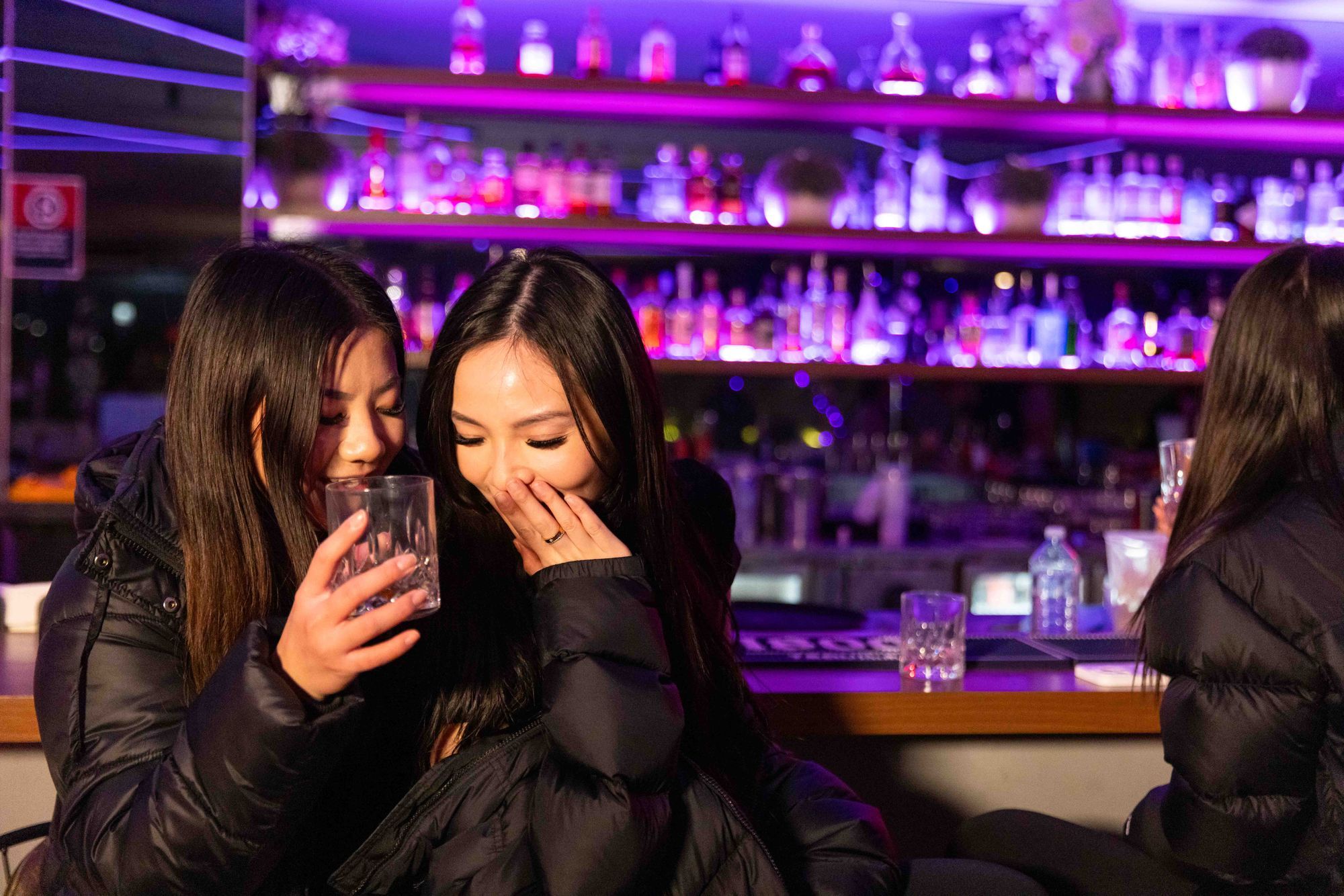 Two women in a bar, laughing and holding drinks, with colourful bottles and dim purple lighting in the background.