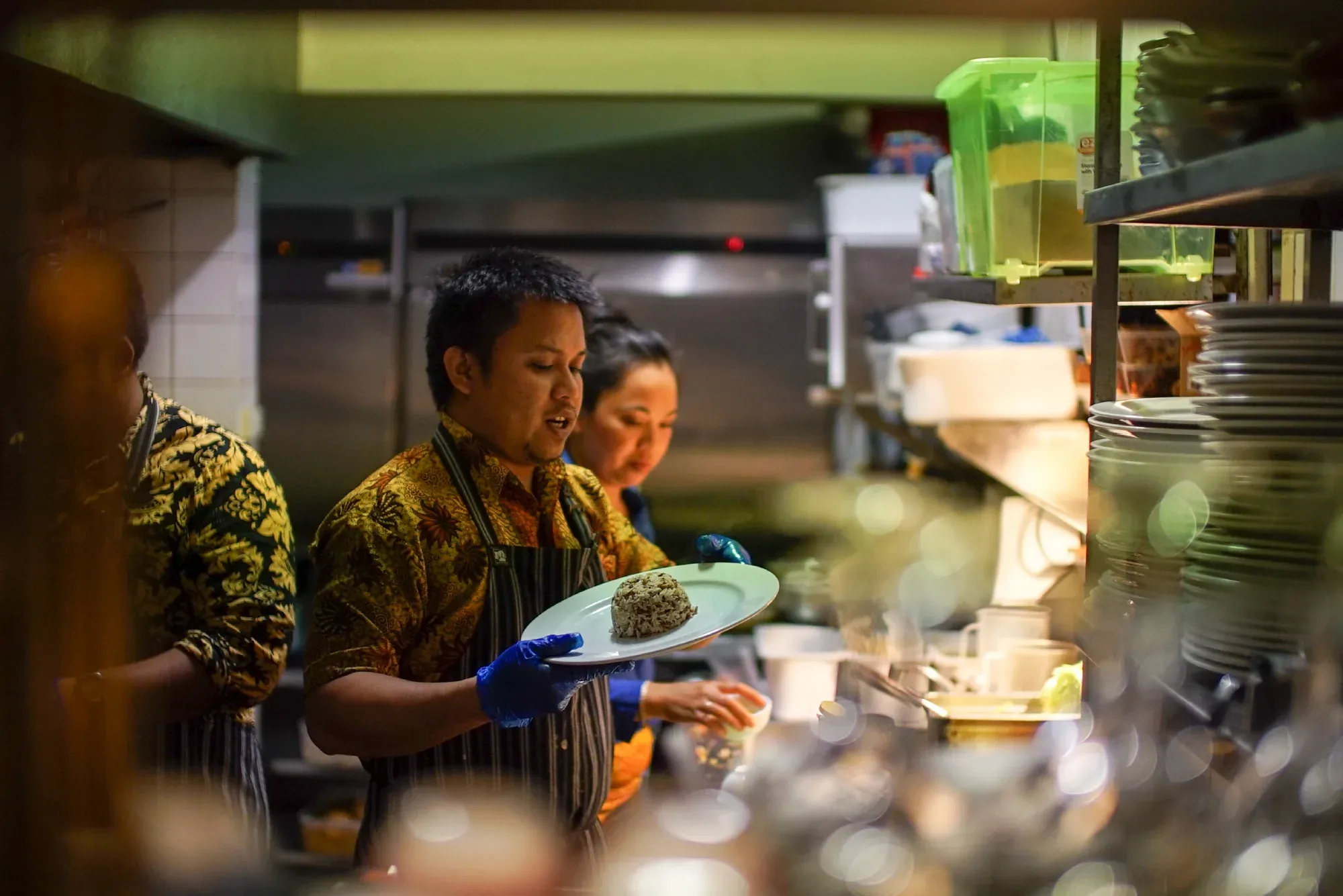 Chef in a busy kitchen, wearing a patterned shirt and apron, holds a plated dish. Another person prepares food in the background.