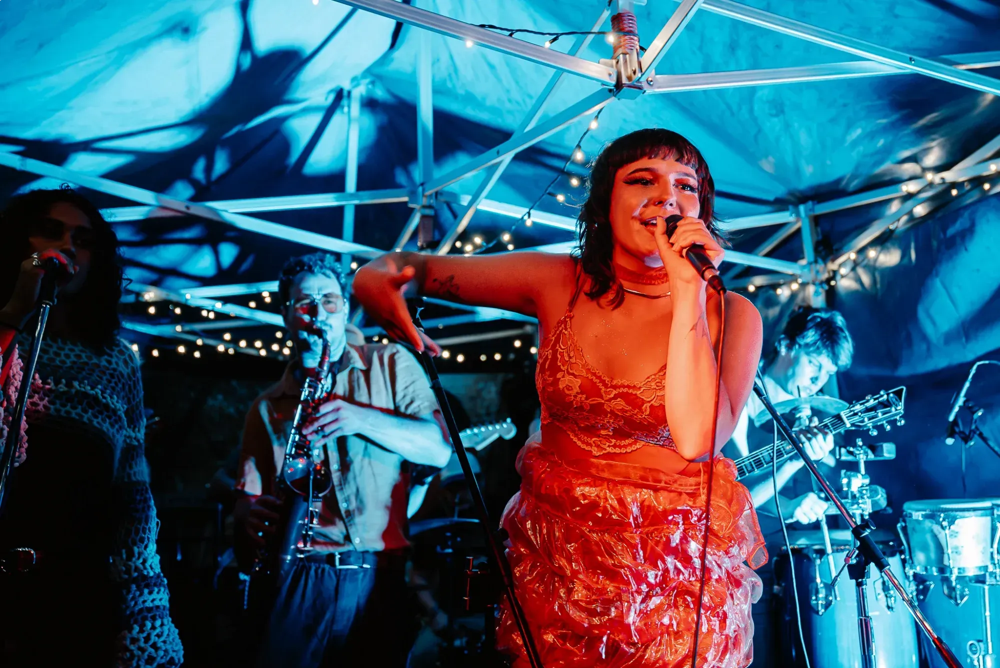 A singer performs passionately under blue lighting, accompanied by a saxophonist and other musicians, with string lights in the background.