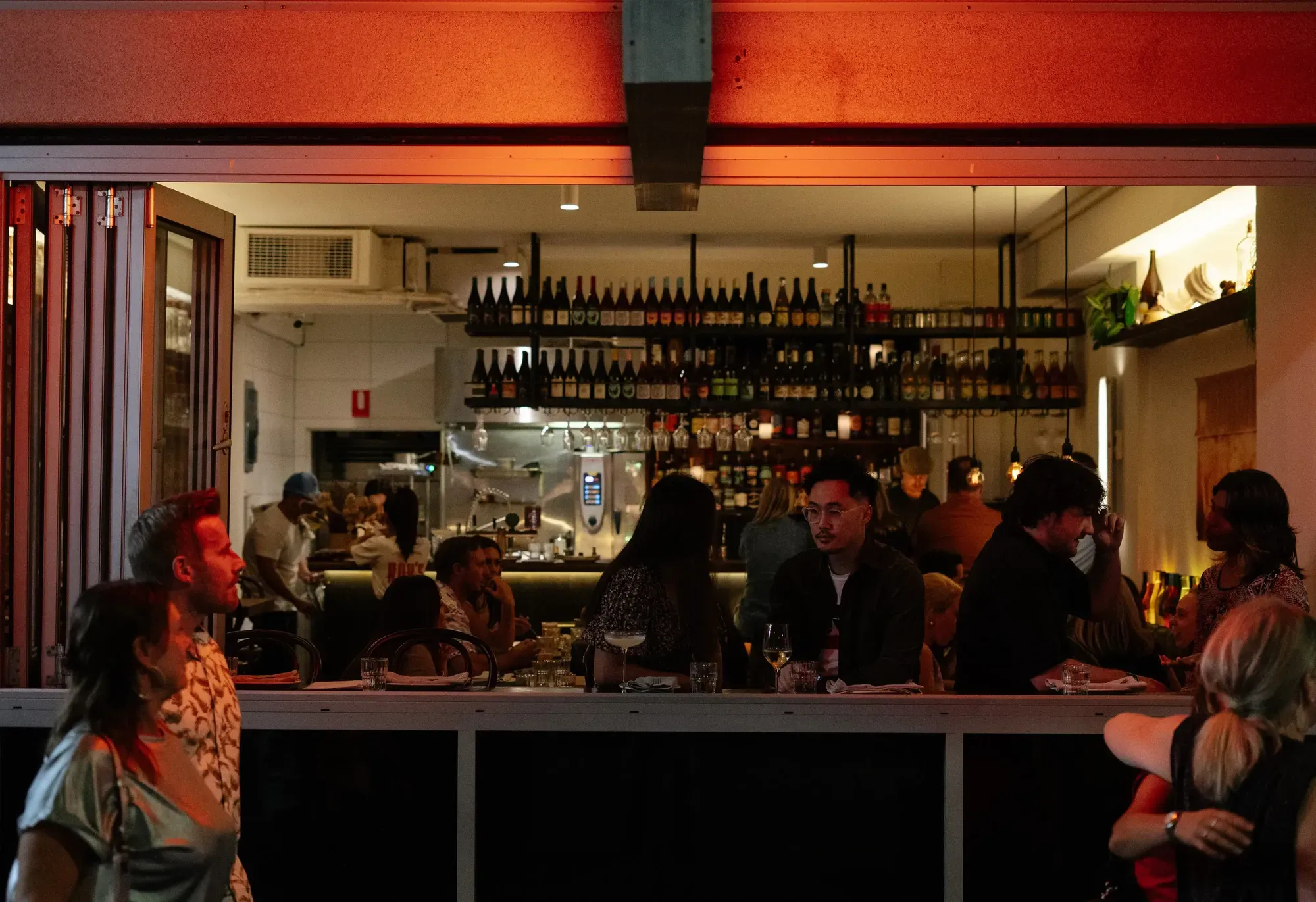 Cosy bar scene with people socializing, dim lighting, and a well-stocked shelf of bottles. Warm ambiance with open kitchen in the background.