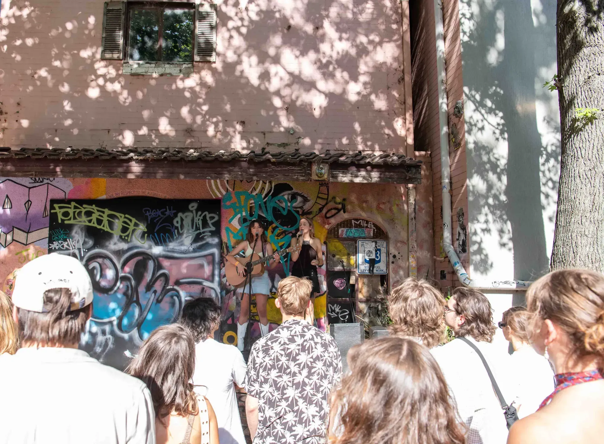 Outdoor performance with two musicians in front of a graffiti-covered wall, surrounded by a small crowd under dappled sunlight.