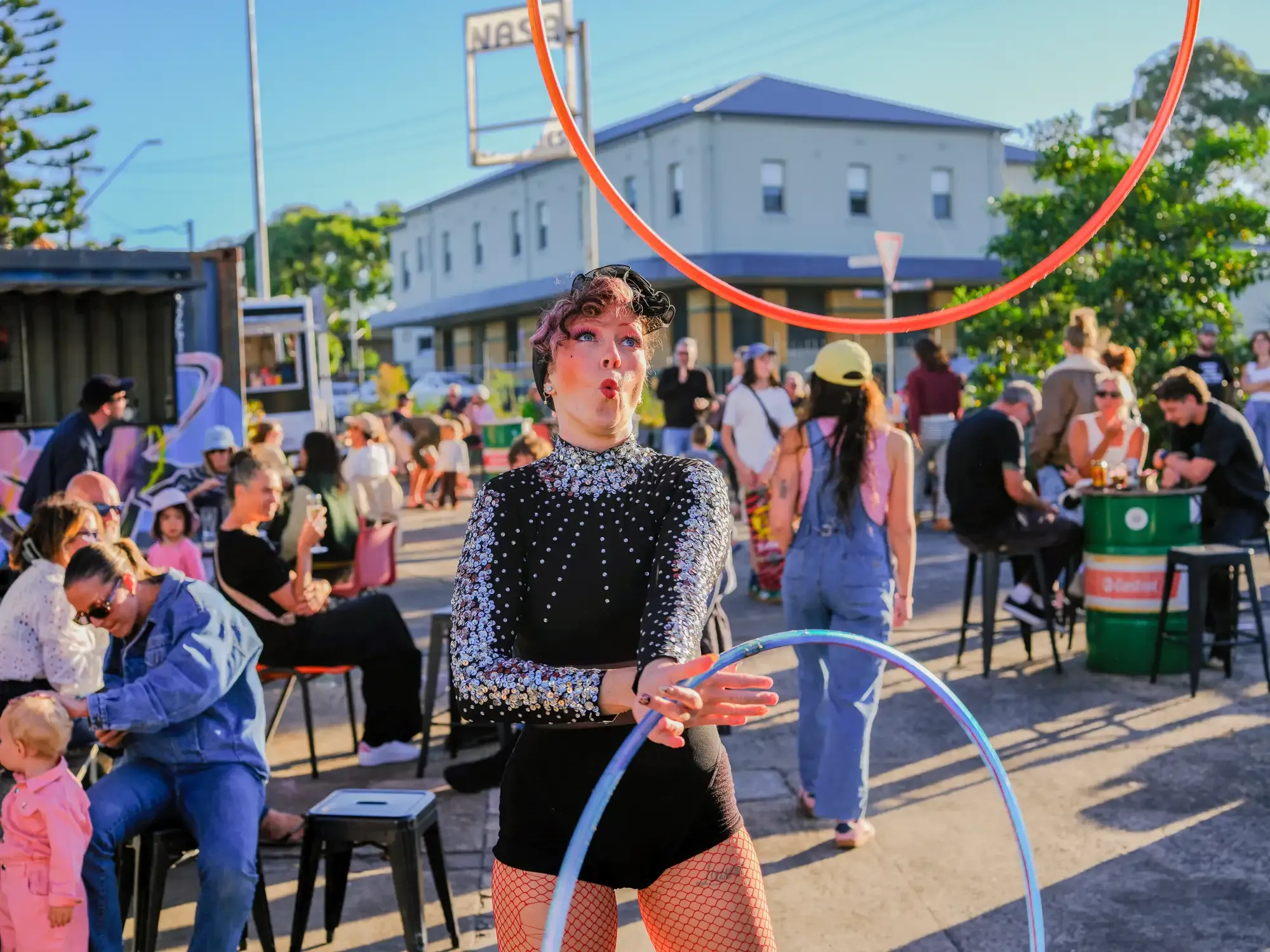 A performer in a sparkly outfit juggles hula hoops at an outdoor event, while people sit and watch against a backdrop of buildings and trees.