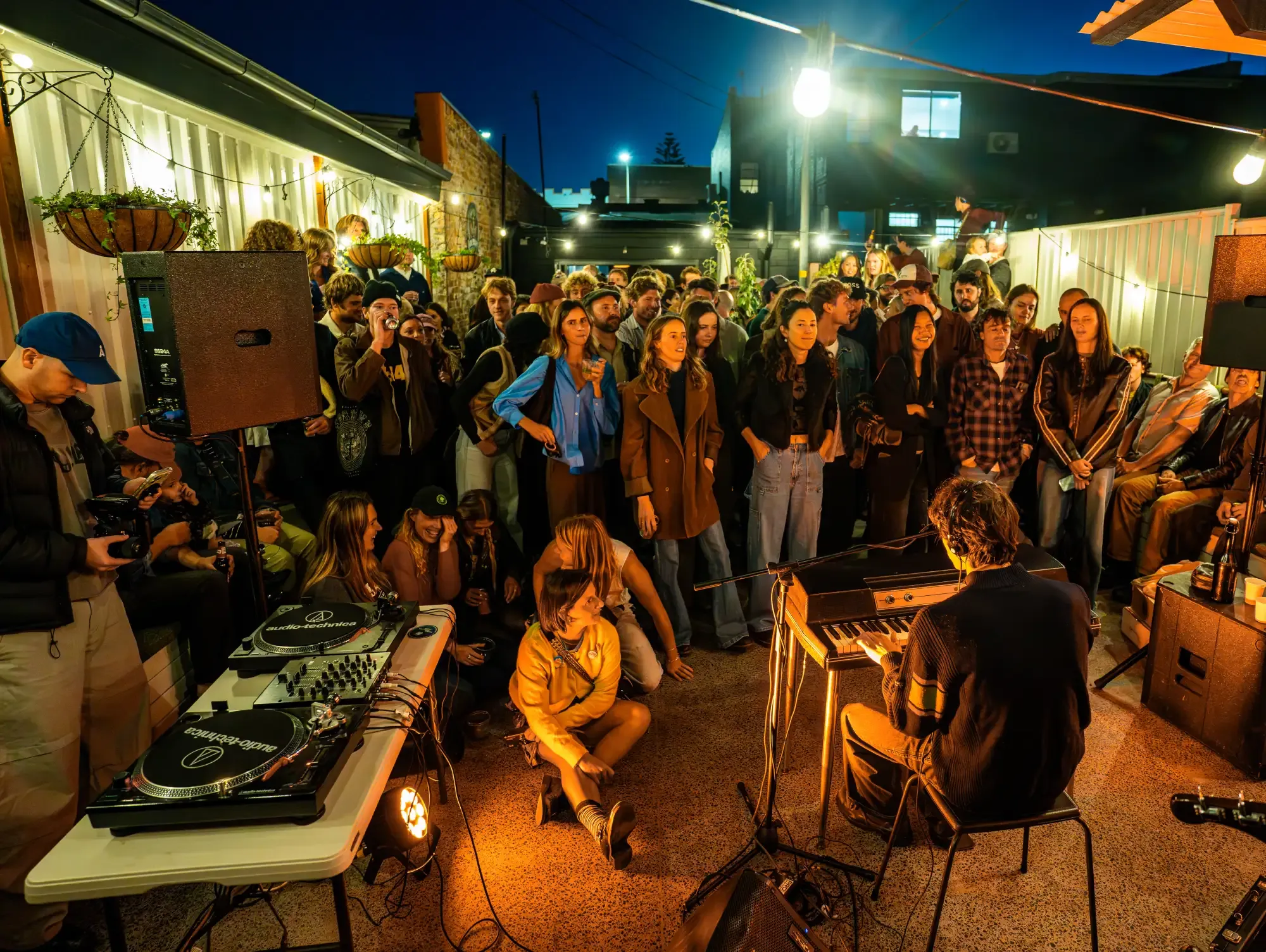 A musician plays a keyboard for an attentive audience at an outdoor night event, with DJ equipment visible and string lights illuminating the scene.