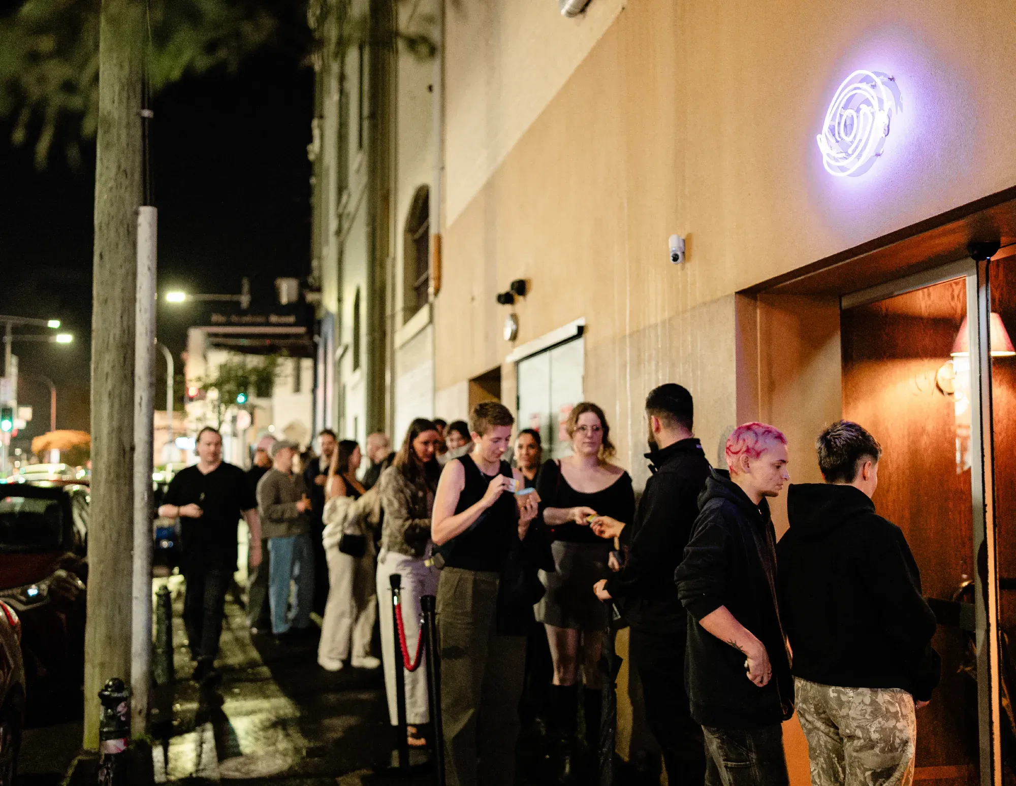 People standing in line outside a dimly lit venue at night, with a blue neon sign above the entrance.
