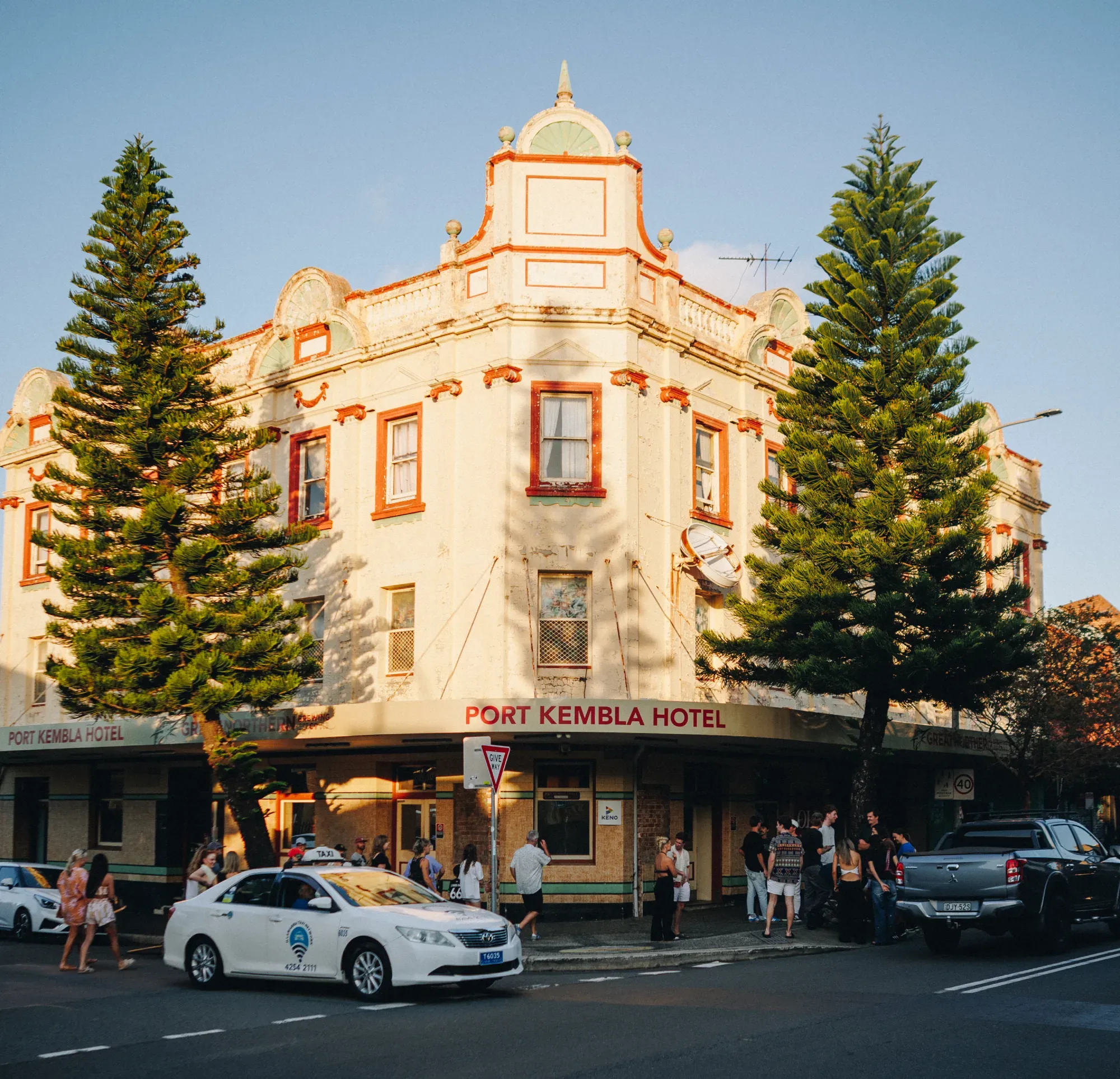 Historic hotel building with trees at corners, "Port Kembla Hotel" sign, and people gathered outside. Cars are moving on the street.