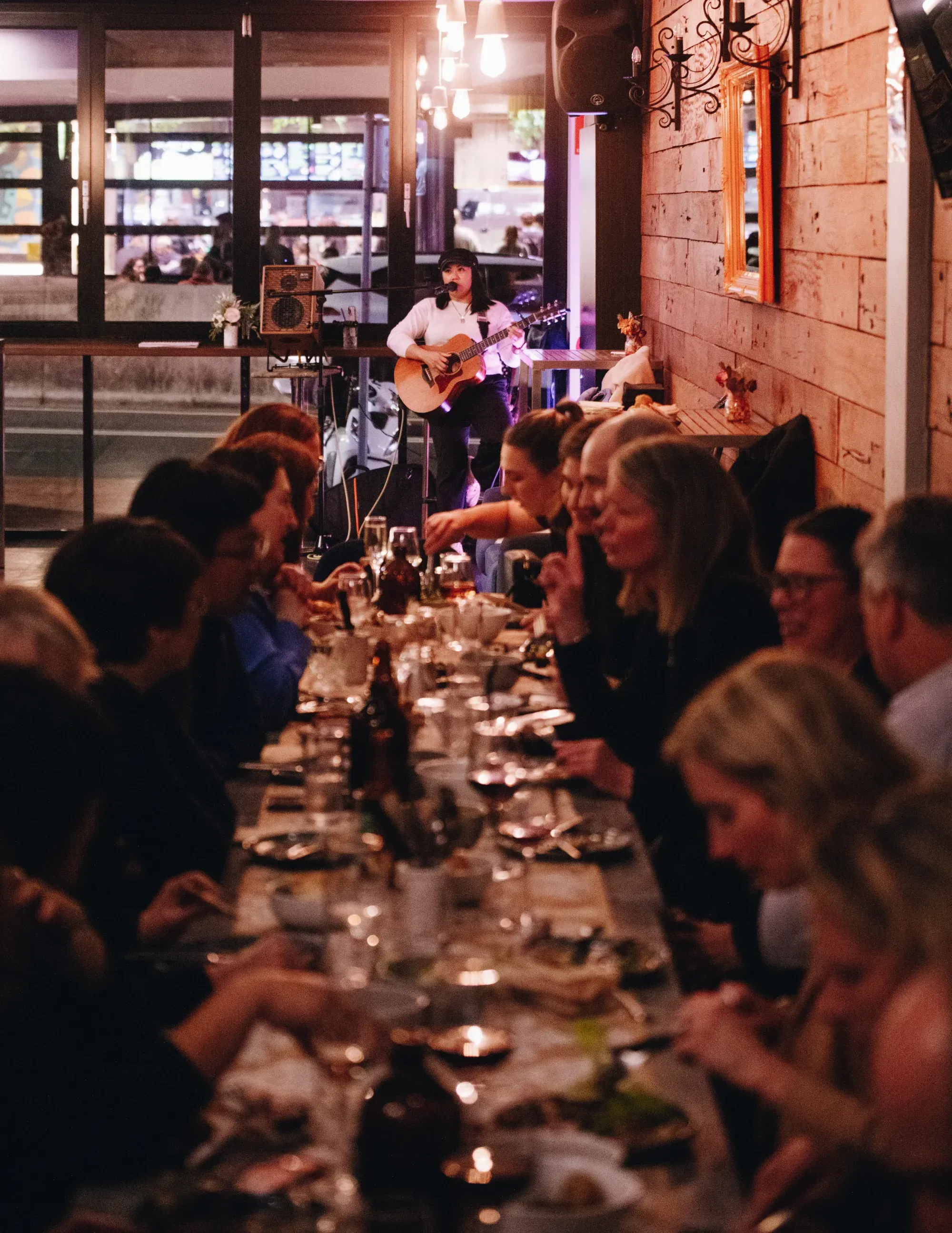 A musician plays guitar at the end of a long dining table filled with people enjoying a meal in a warmly lit restaurant.