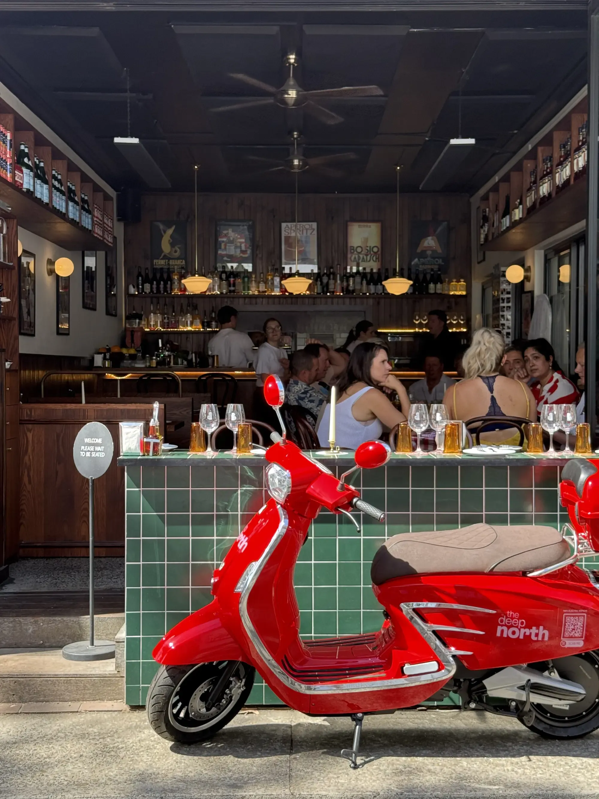 A bright red scooter is parked outside a lively cafe. Patrons inside are seated at a green-tiled bar, enjoying drinks and conversation.