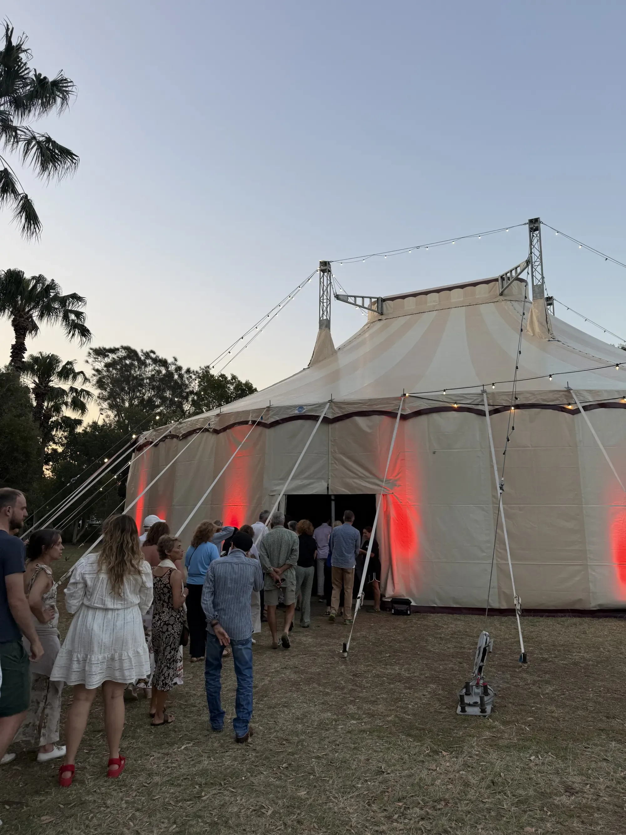 People lining up to enter a large, illuminated circus tent at dusk, with strings of lights and palm trees in the background.