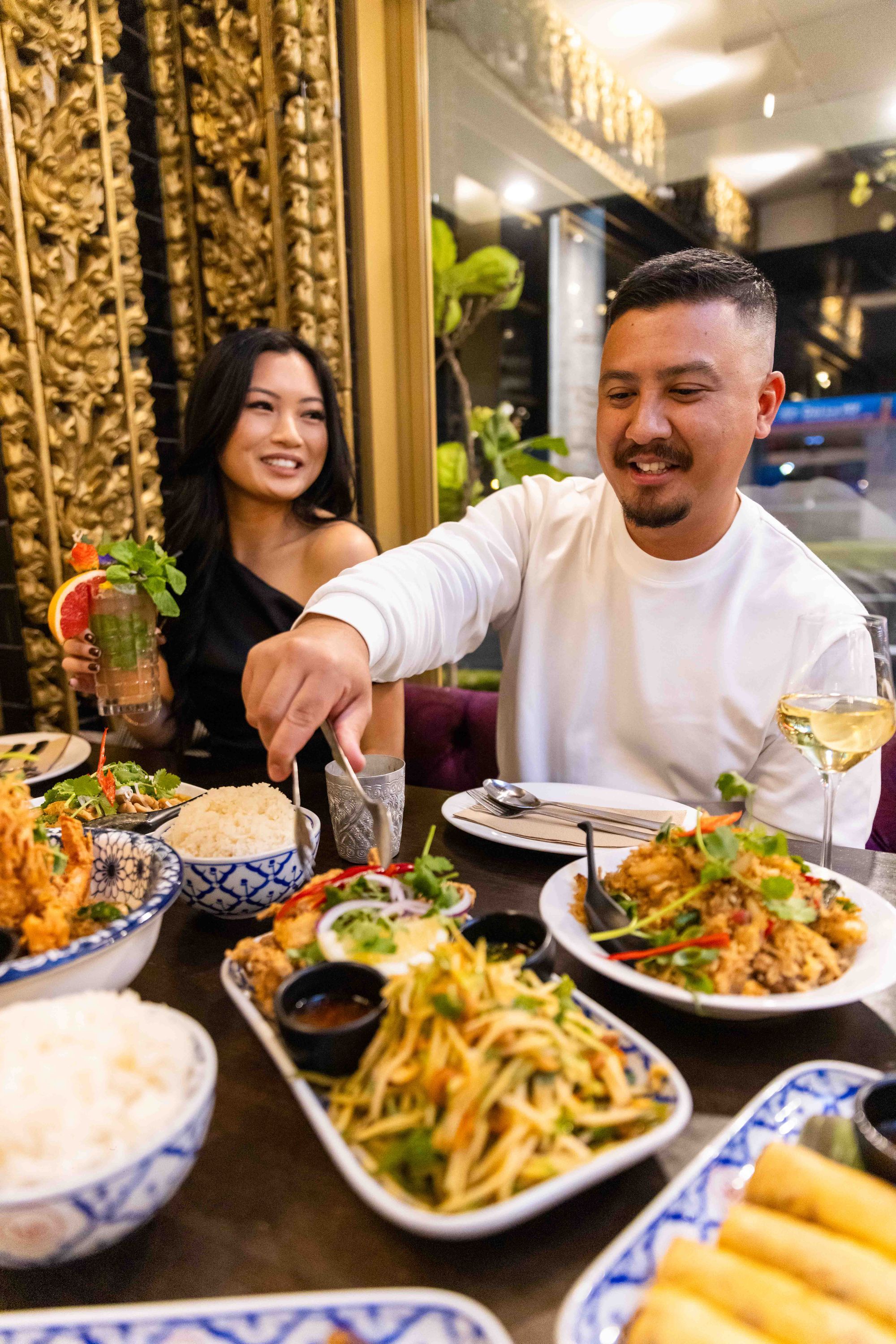 A couple enjoys a meal at a restaurant, with a table full of colorful dishes and drinks. The man serves food while the woman smiles.