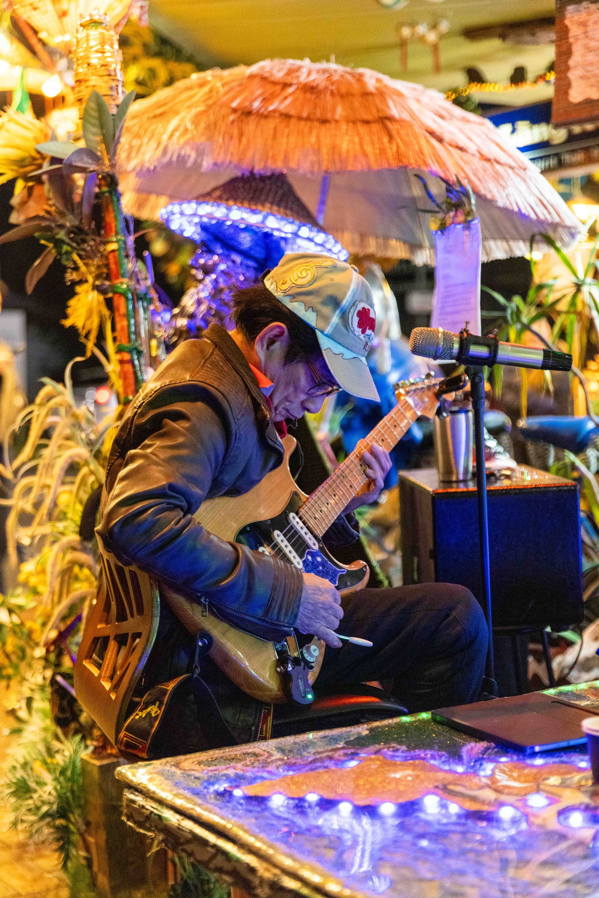 Musician in a jacket and cap plays an electric guitar under a colorful umbrella, surrounded by vibrant decor and plants.