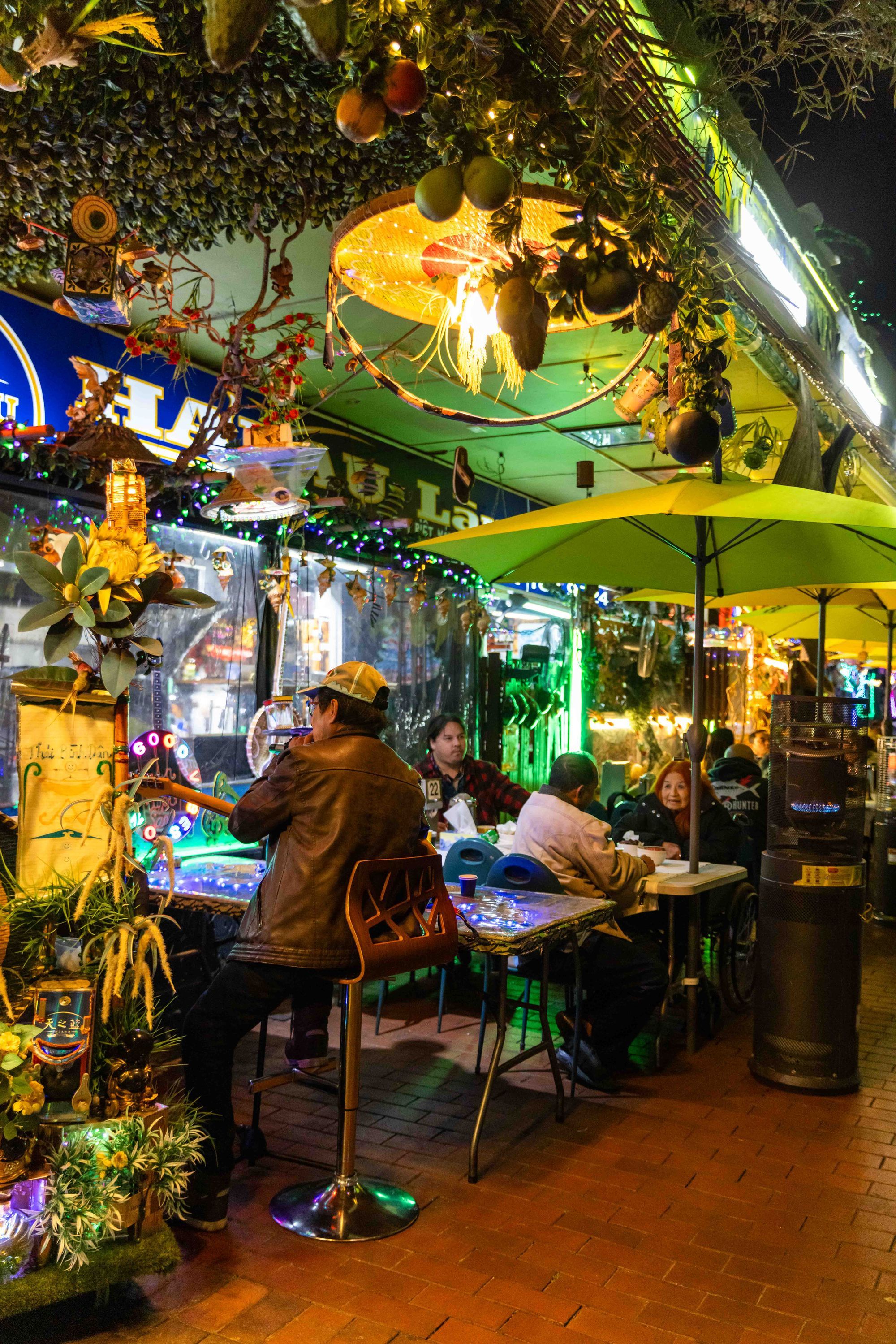 Outdoor restaurant with people dining under green umbrellas, surrounded by colorful lights and decorations, creating a festive atmosphere.