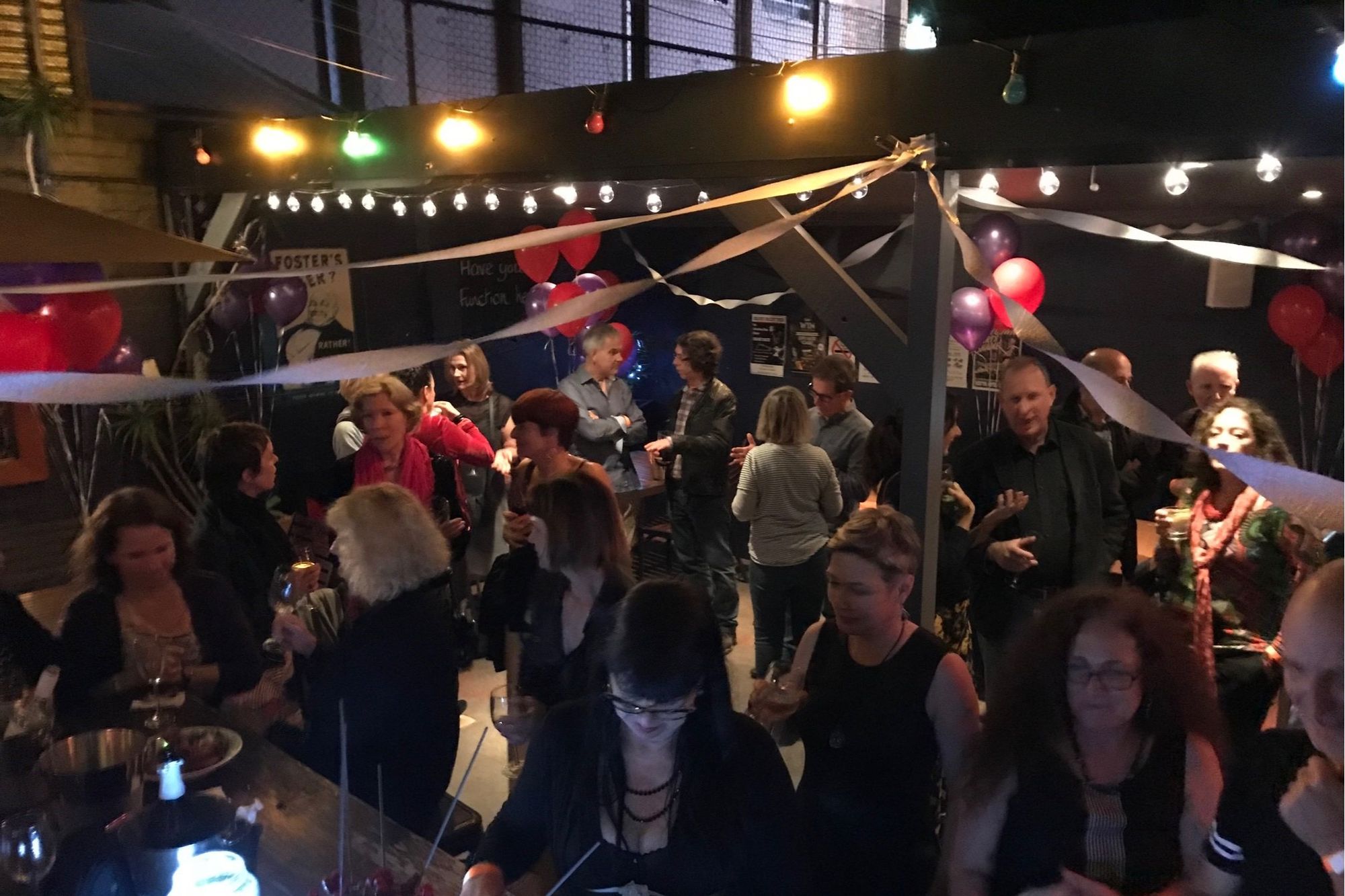 A lively party scene with people socializing under string lights and balloons, surrounded by festive decorations and a relaxed indoor-outdoor atmosphere.