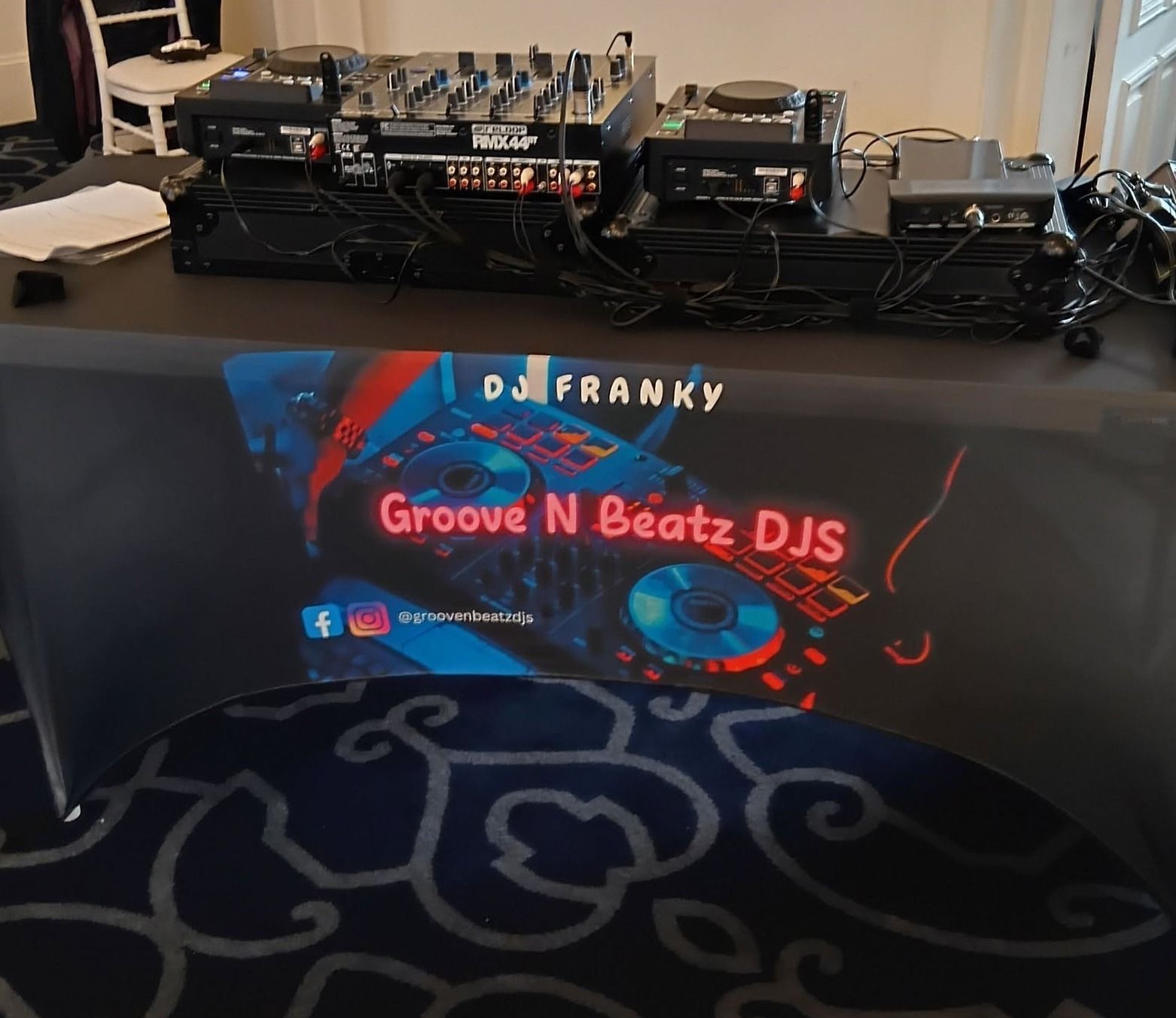A DJ setup with mixers and sound equipment on a table draped with a cloth showing "DJ Franky" and "Groove N Beatz DJS" with social media symbols.