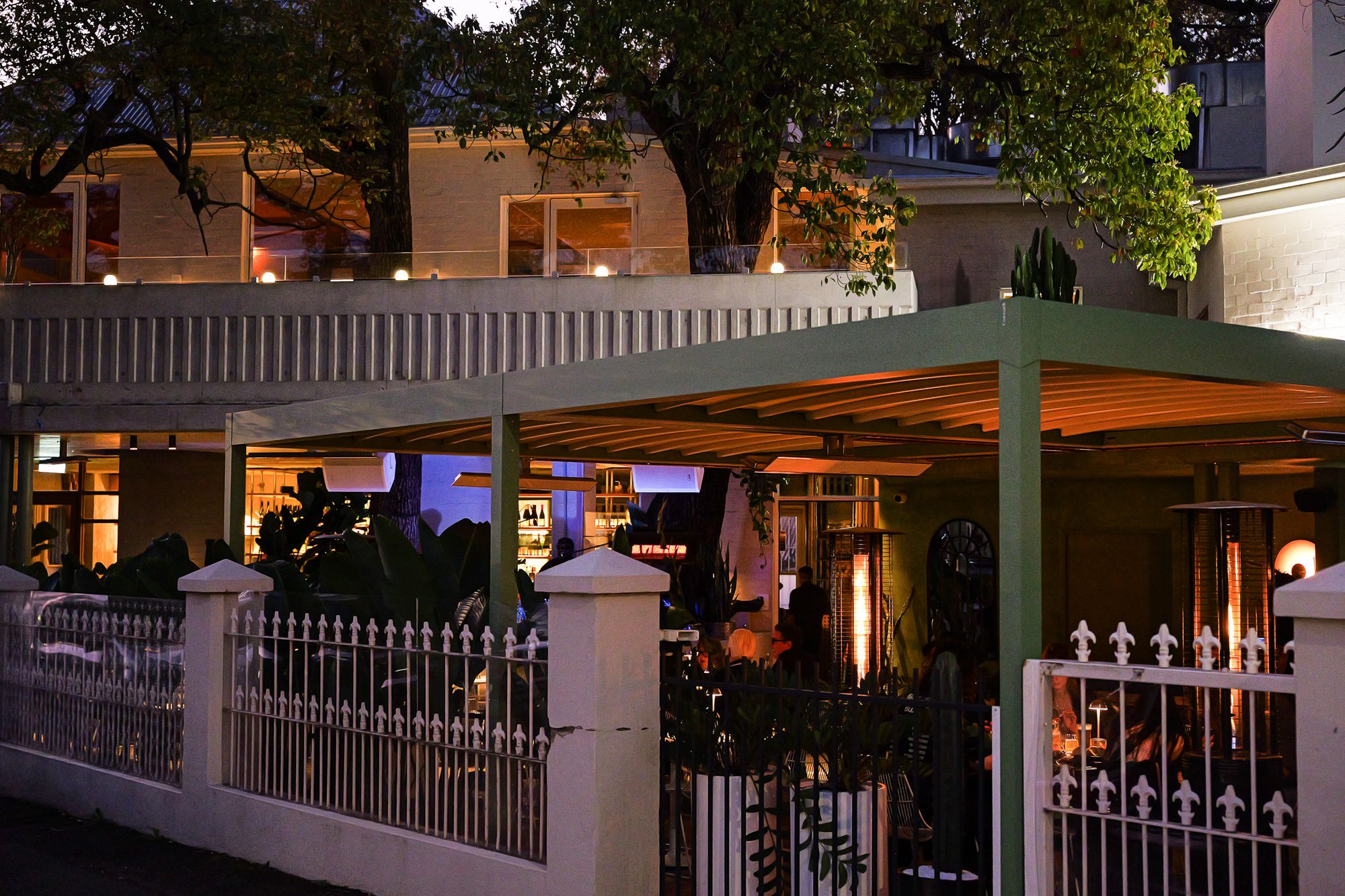Outdoor restaurant patio at dusk with green pergola, ambient lighting, and lush greenery, set behind a decorative iron fence.
