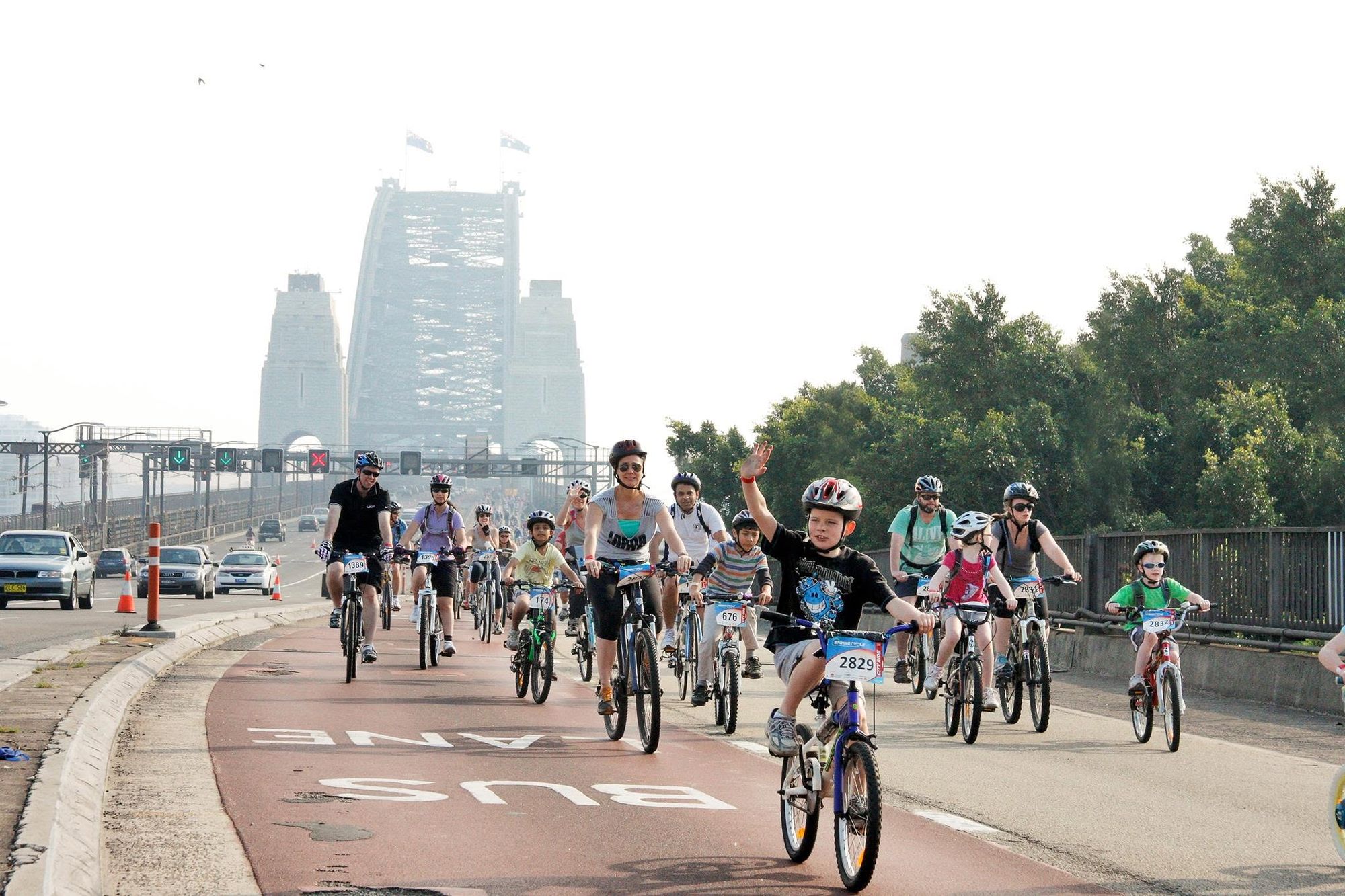 A group of cyclists, including children and adults, ride on a roadway near a large bridge, enjoying a sunny day with helmets on.