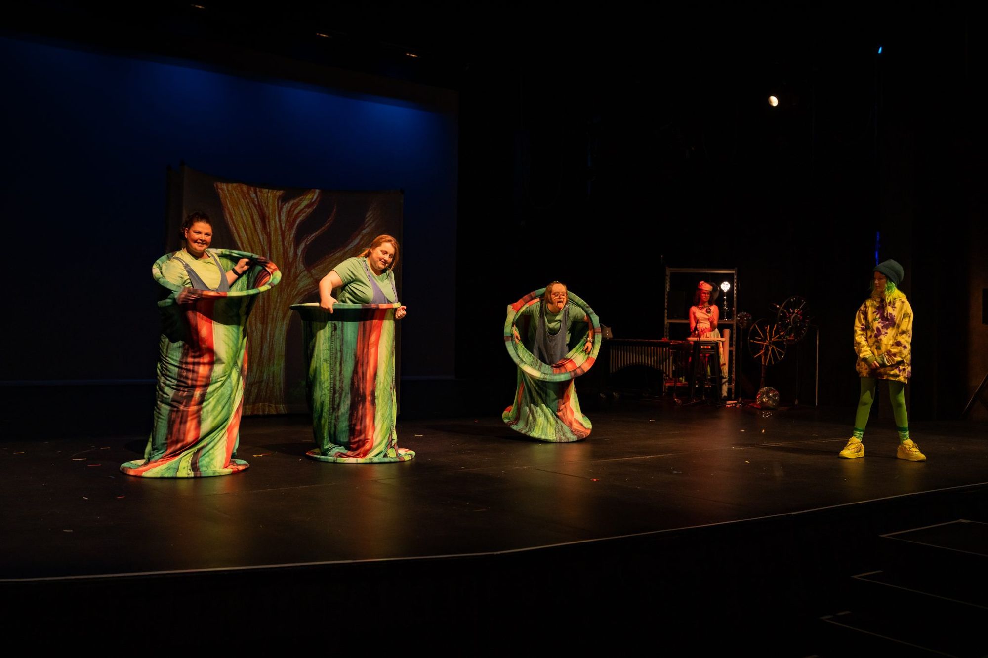 Performers in colourful costumes stand on stage, with one person in a yellow outfit to the right, under dim lighting.