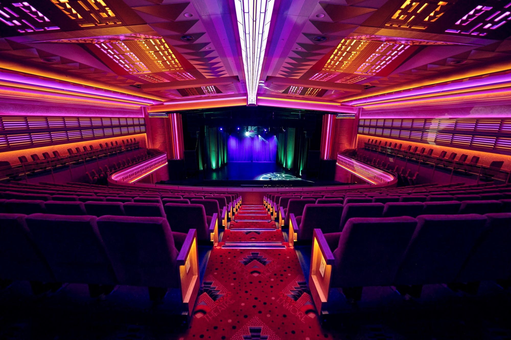 Vibrant theatre with illuminated stage and rows of red seats. Neon lights create a colorful ambiance throughout the interior.