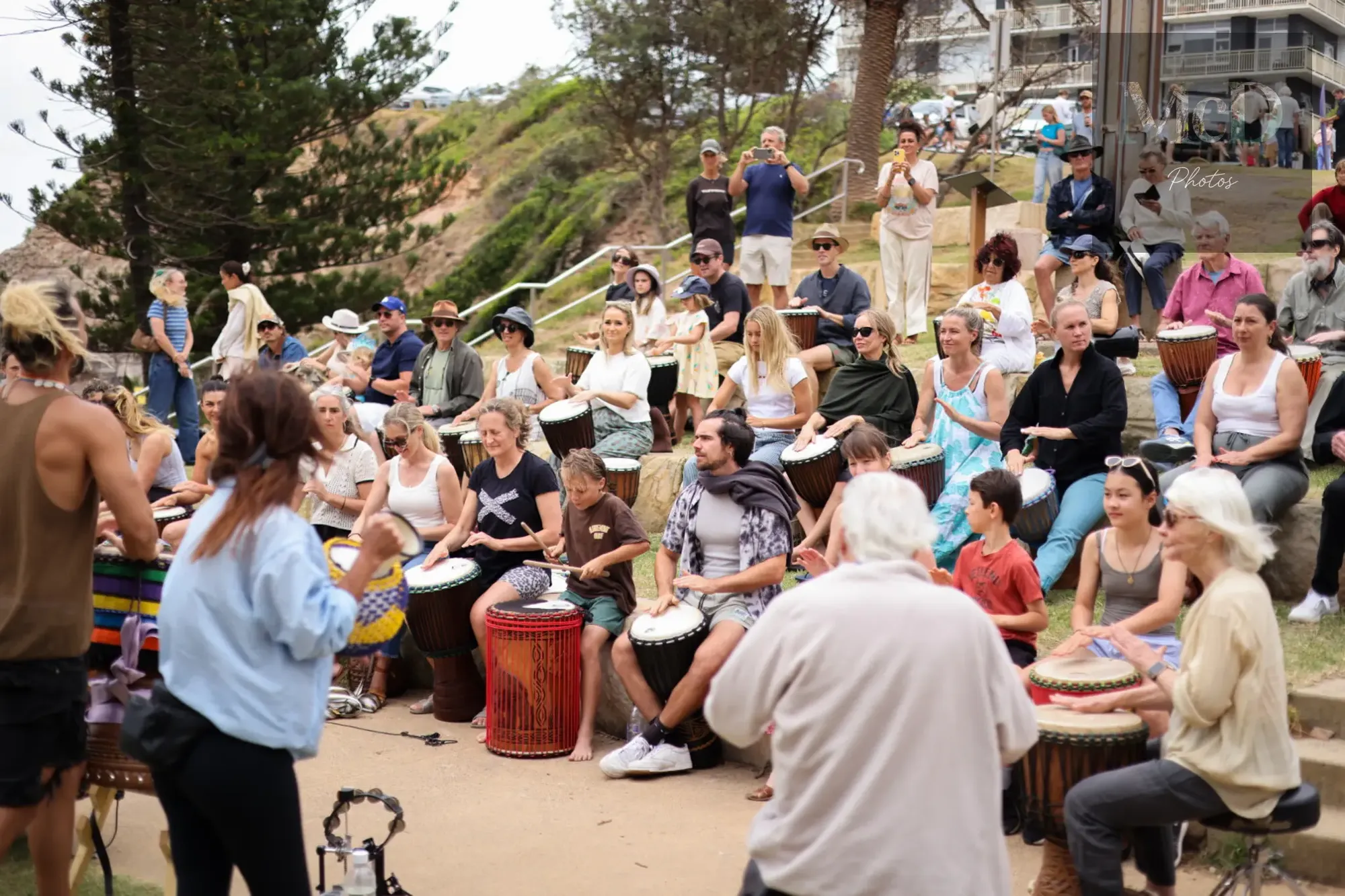 A diverse group of people engage in a lively outdoor drumming circle, surrounded by trees and an audience on a grassy slope.
