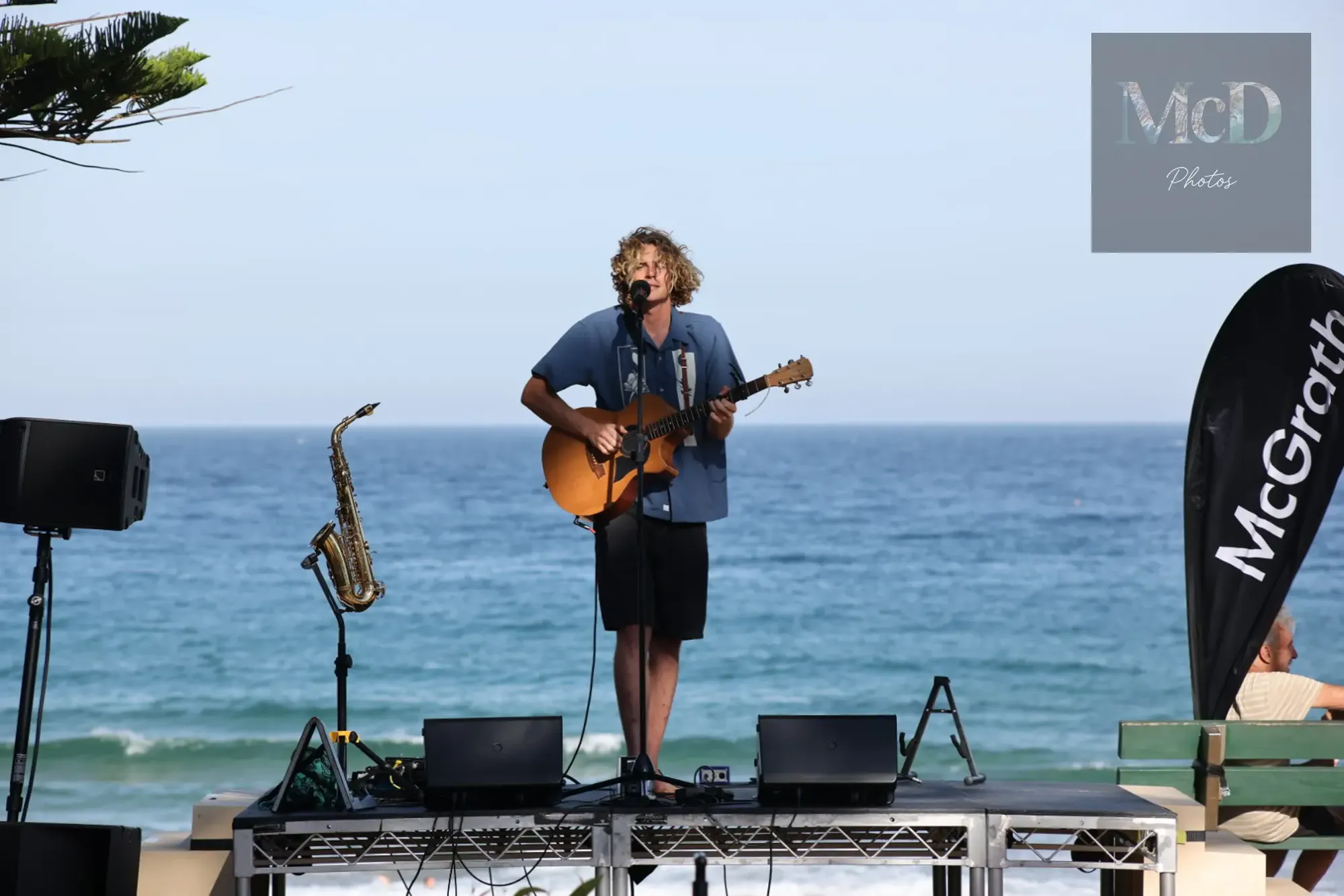 Musician playing guitar on a platform by the sea, with a saxophone beside him and McGrath signage on a flag.
