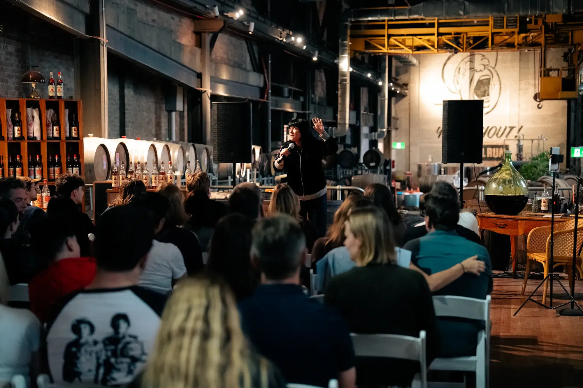 A woman speaks into a microphone in a distillery setting, with an audience seated in front. Industrial decor and barrels are visible in the background.
