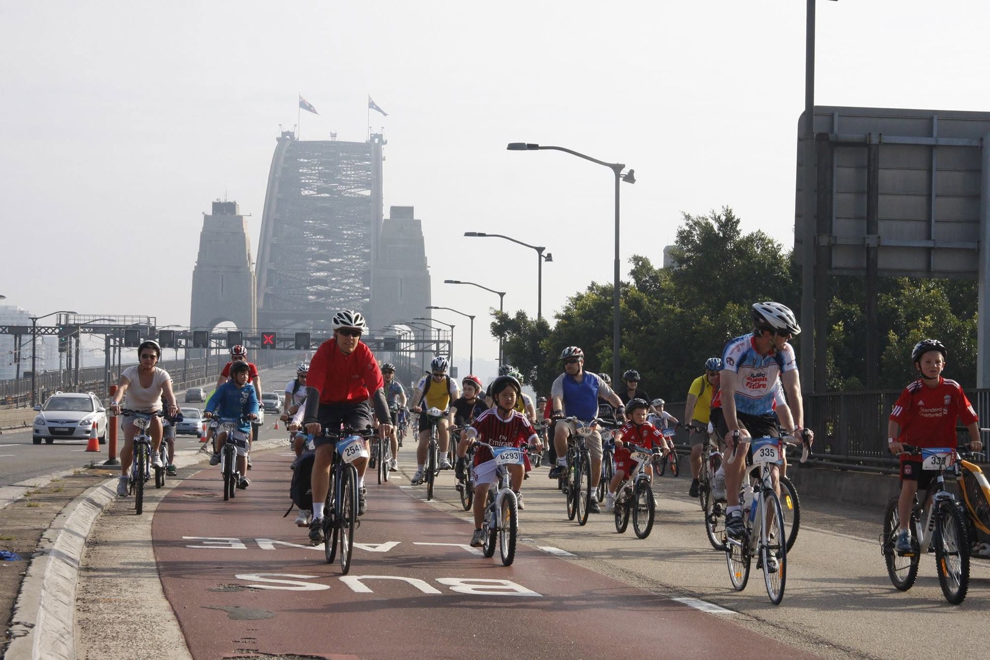 A group of cyclists, including children, ride along a designated bike lane on a bridge, with the bridge structure visible in the background.