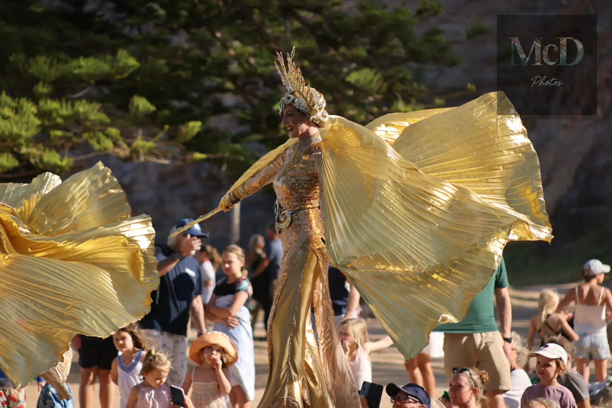Performer on stilts in a gold costume with flowing wings, surrounded by a crowd at an outdoor event; trees in the background.