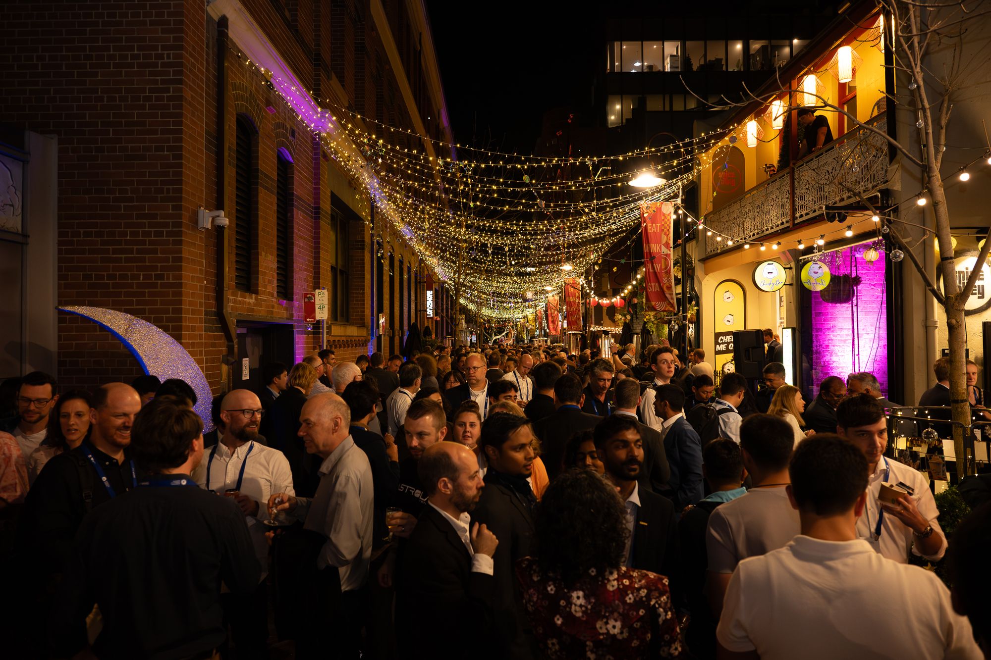 Crowded nighttime street festival under a canopy of twinkling string lights, people mingling between brick buildings and lit shopfronts.