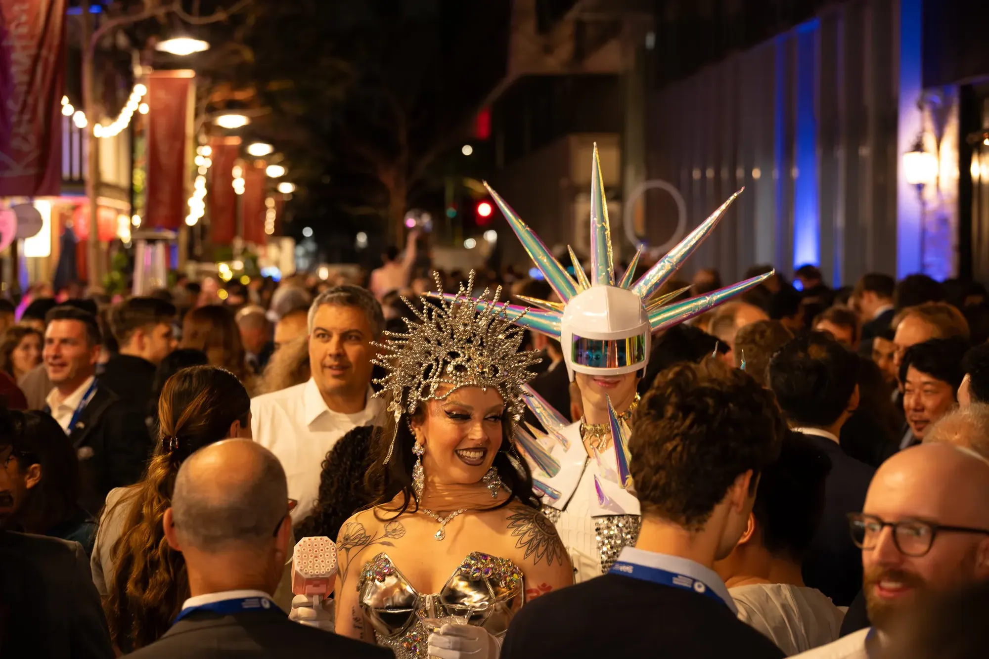 A lively street crowd, with a person in a spiky headdress and another in a star helmet, both in elaborate costumes, engaging with attendees.