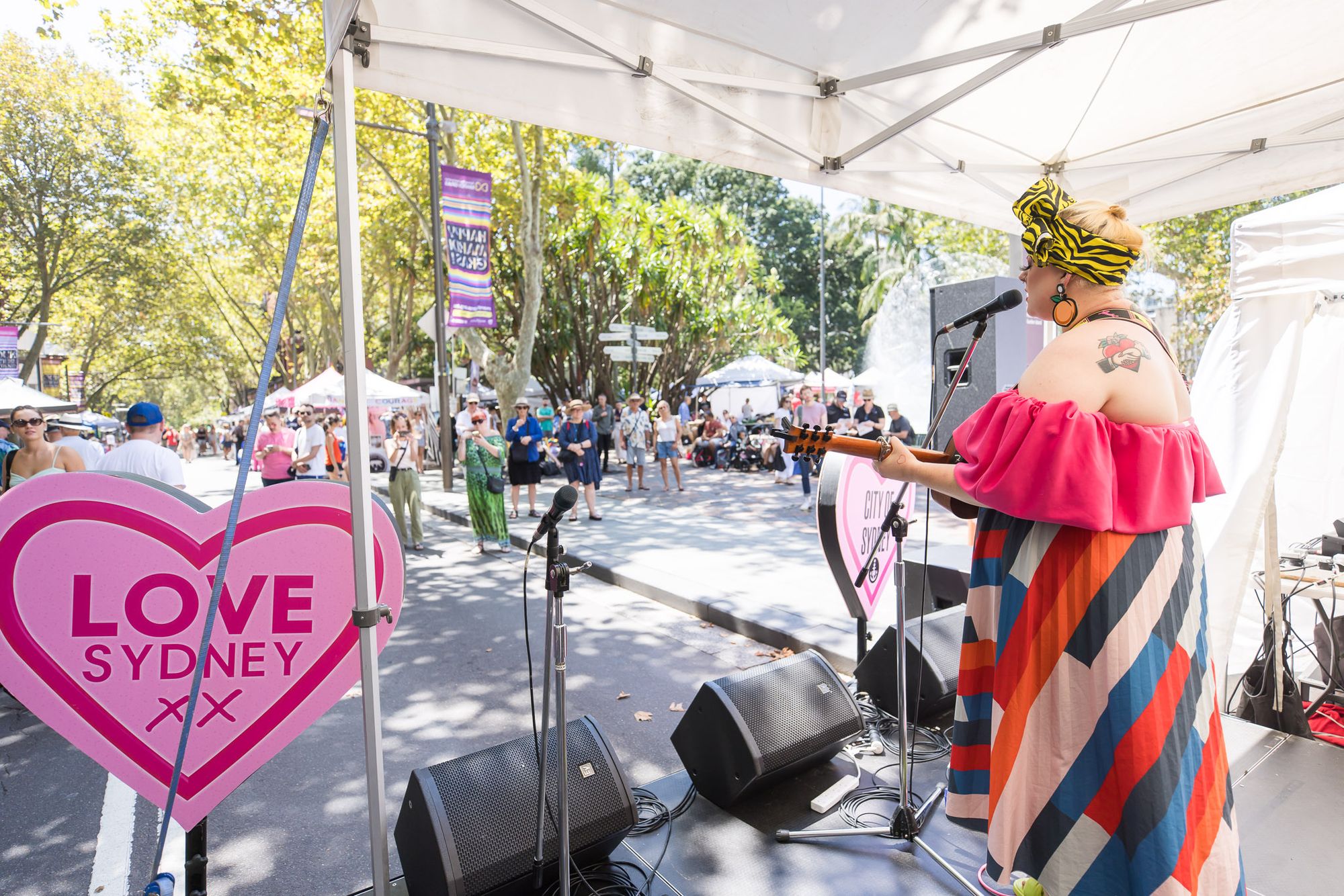 A performer plays the guitar and sings on a small stage on the street. A crowd of people in the background look on.