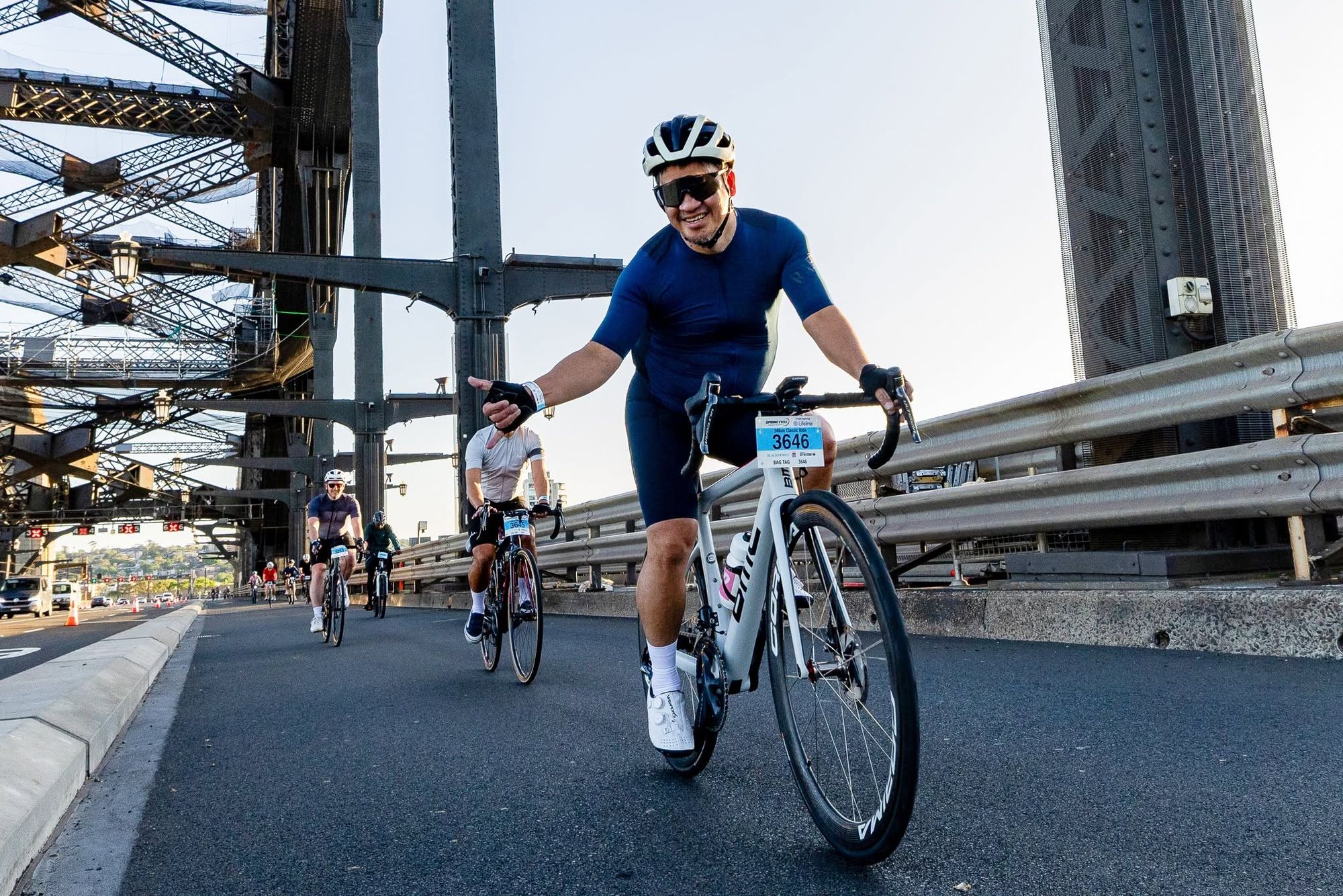 Cyclists riding across a bridge. The leading cyclist in a blue outfit smiles and gestures while others follow. Clear sky and bridge structure visible.