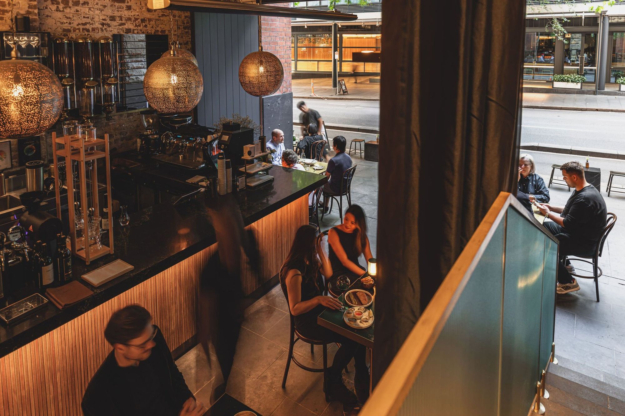 Cozy café interior with dim lighting and patrons seated at tables, some enjoying drinks by the window with a view of the street outside.