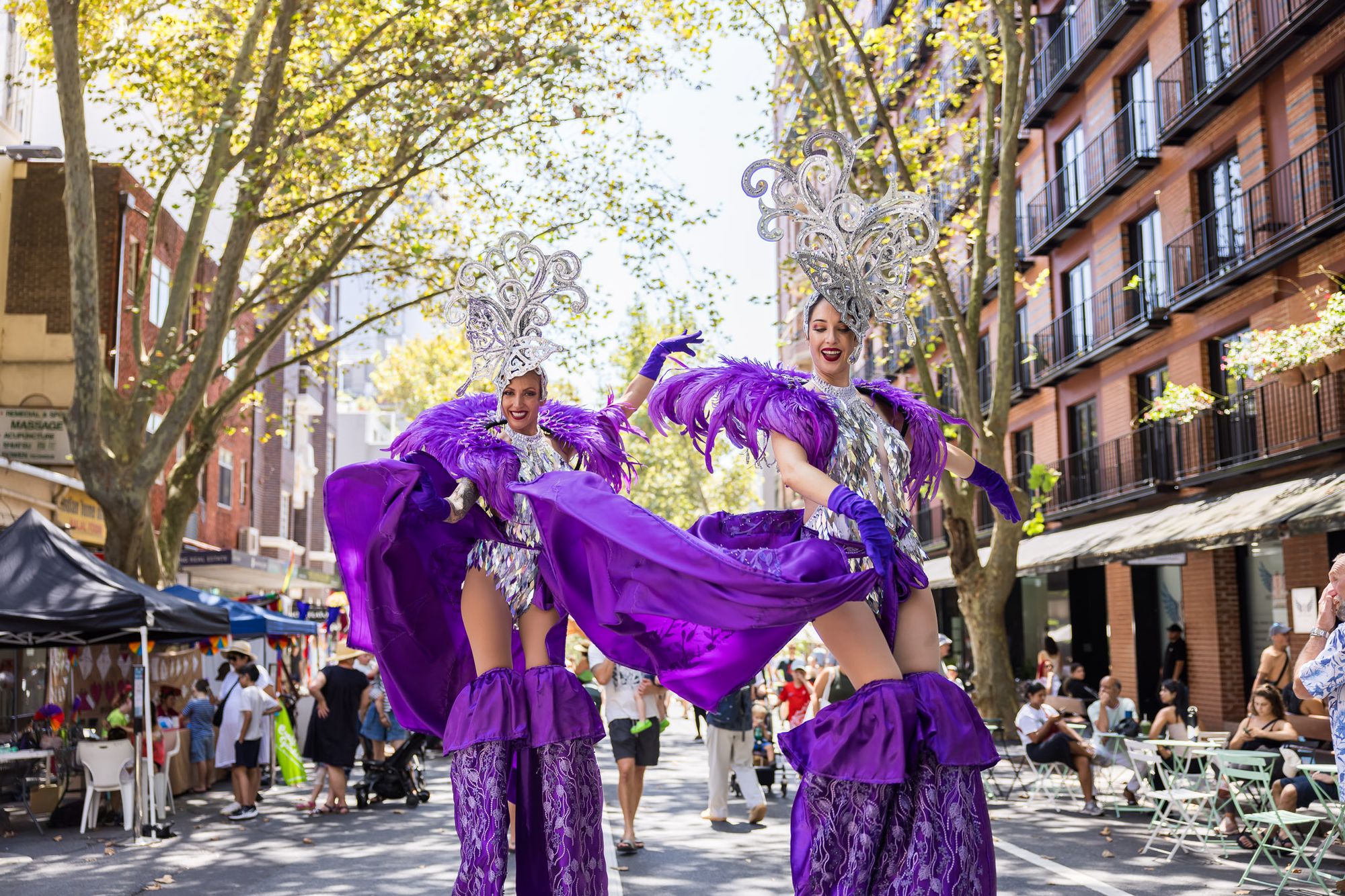 Two performers in purple with elaborate silver head dresses stand on stilts in the middle of a high street festival